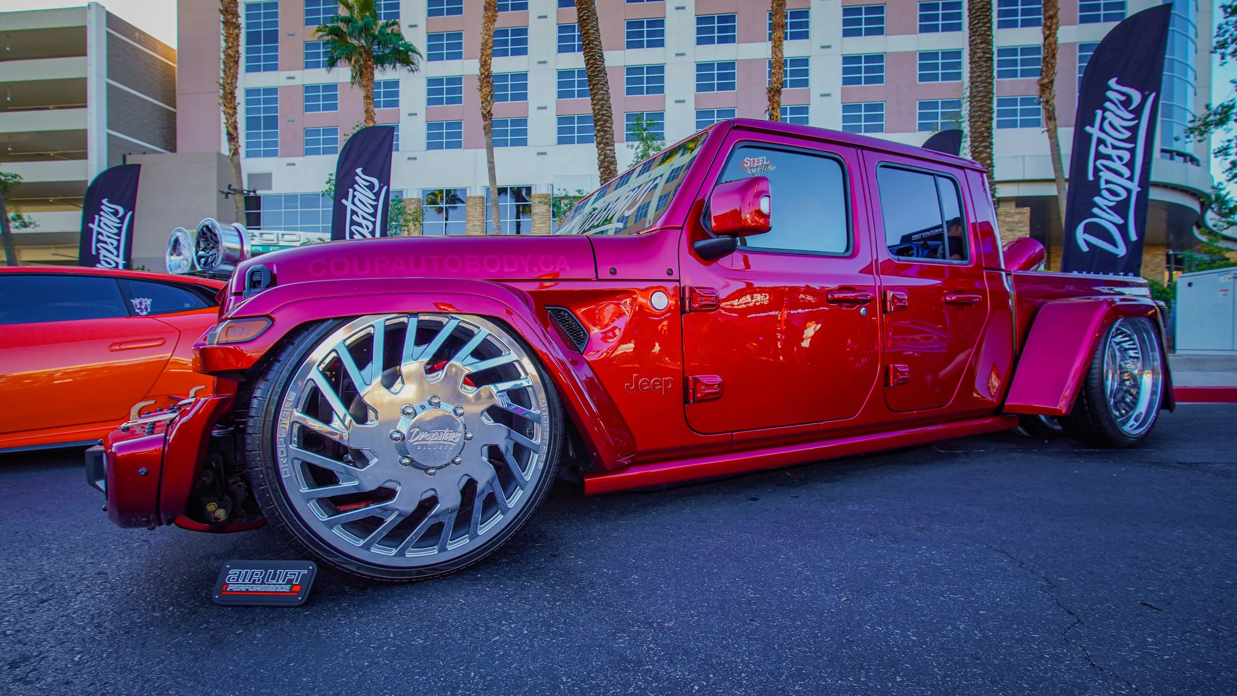 Red customized Jeep truck with large chrome wheels and lowered suspension parked outdoors at a car show.