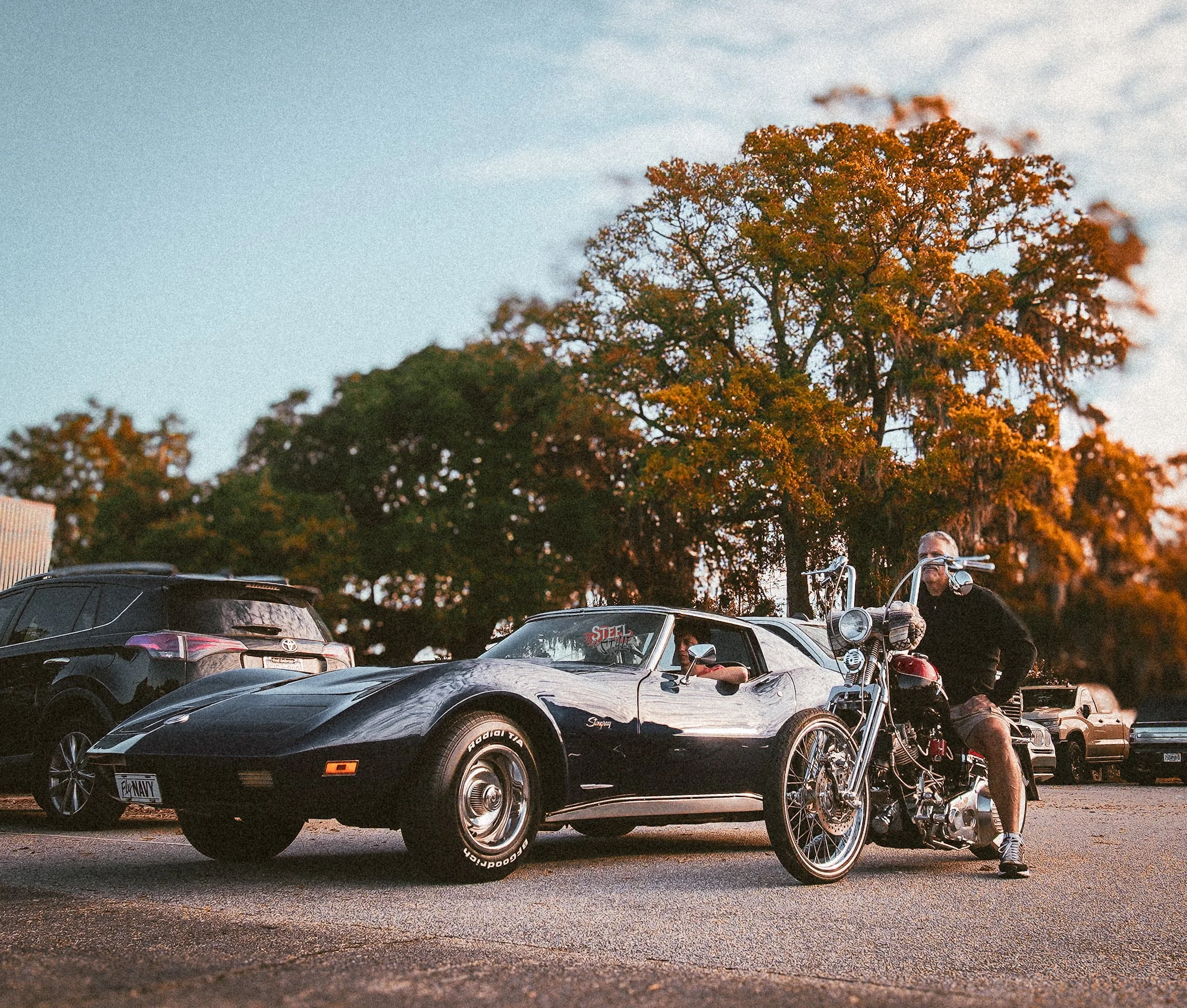 A man sitting on a motorcycle parked next to a black sports car, with trees and other parked cars in the background during sunset.