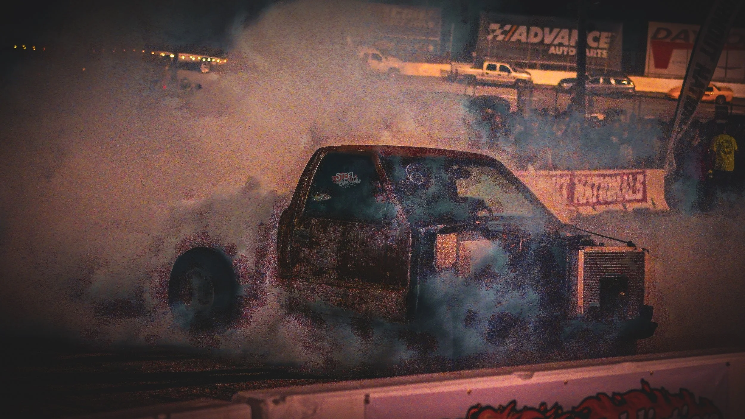 A drag racing car emits smoke during a nighttime race at a drag strip, with spectators and parked cars visible in the background.