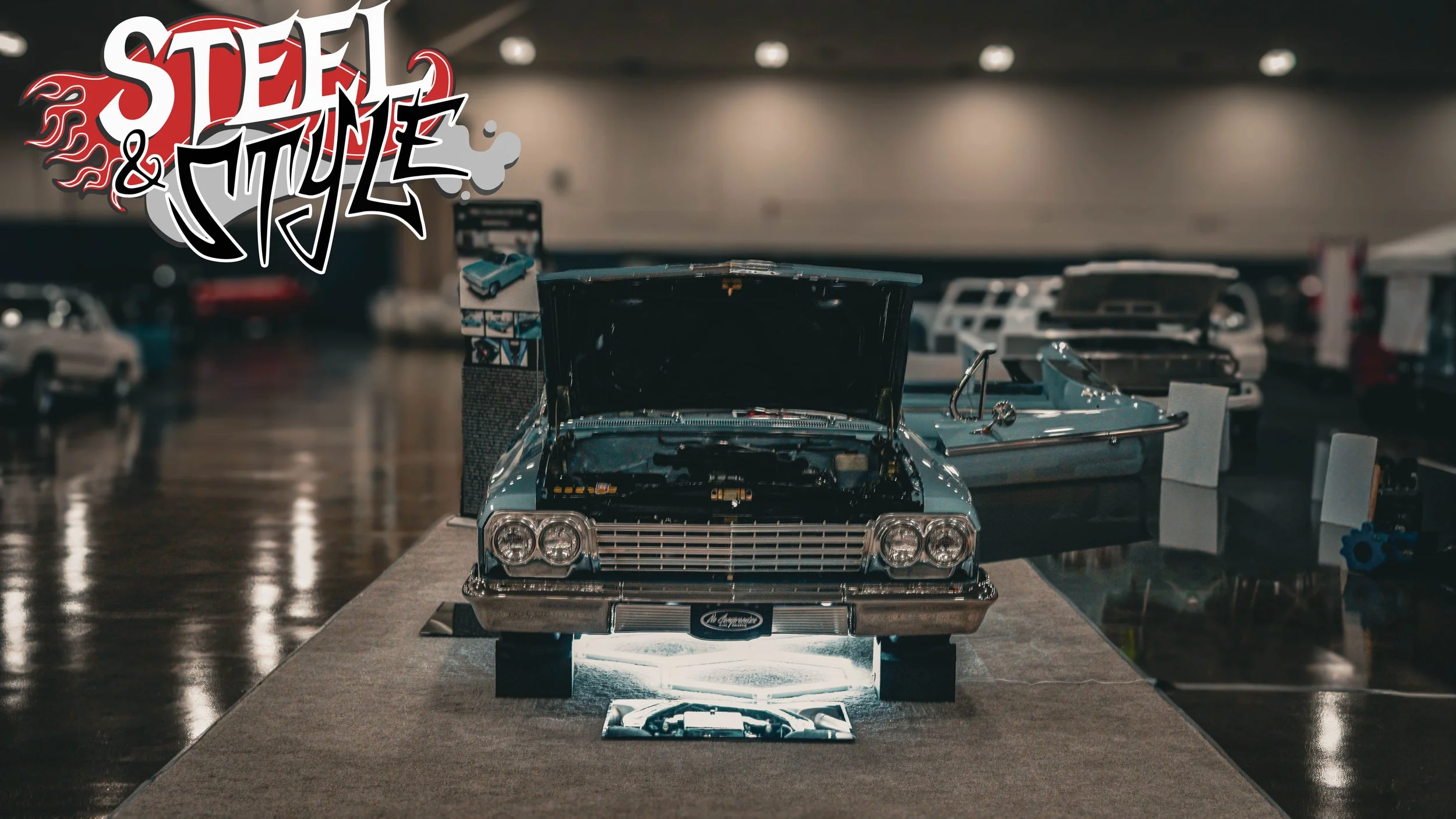 Model of a vintage blue car with its hood open on display at an indoor exhibition, with other vintage cars visible in the background.