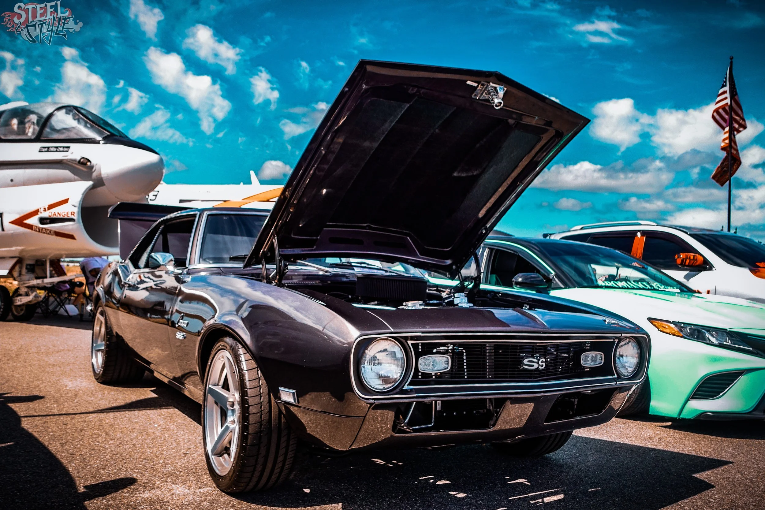 A classic black Chevrolet Corvette with its hood open at an outdoor car show, with other cars and an aircraft in the background under a partly cloudy sky.