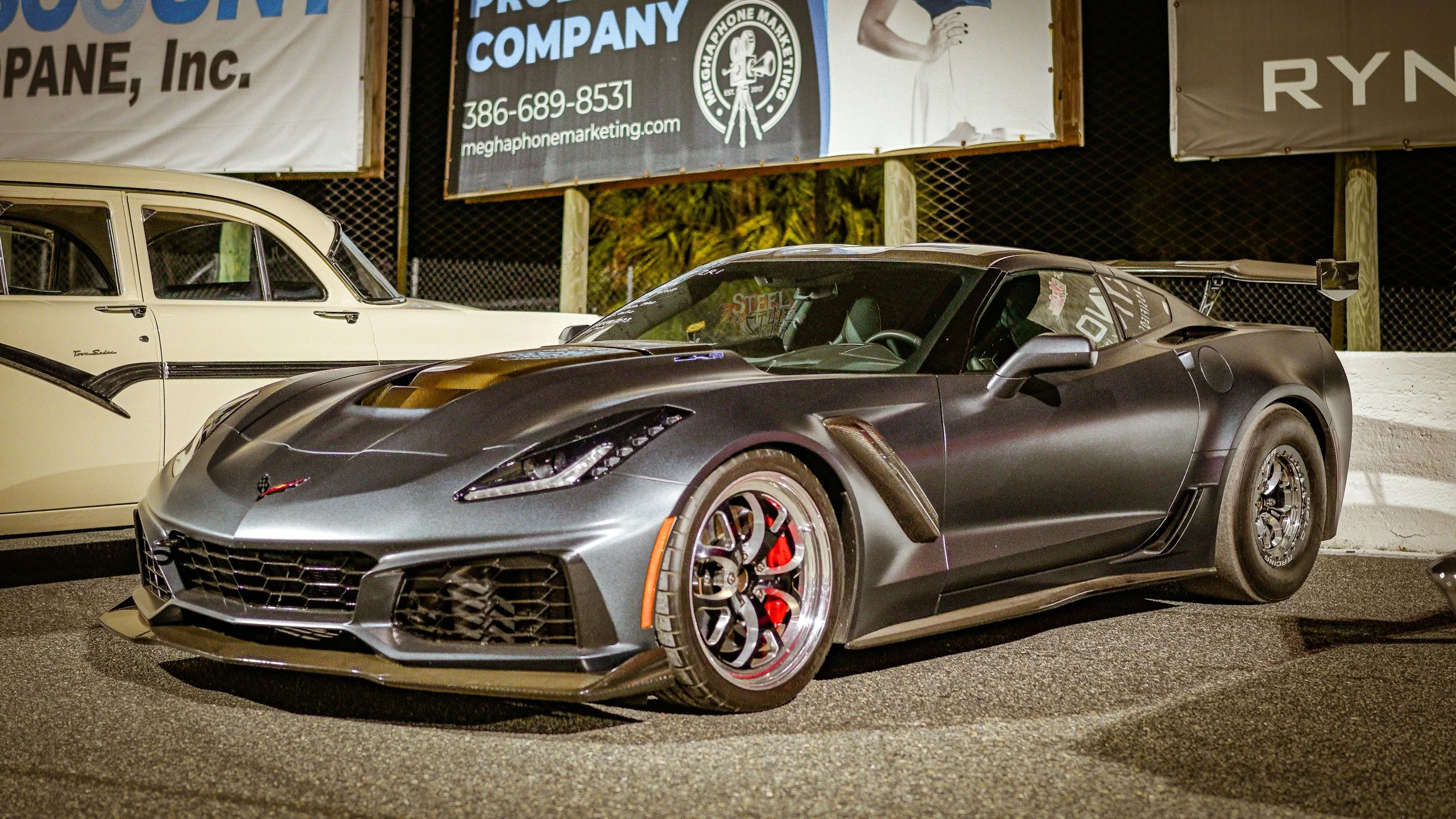 A gray Chevrolet Corvette Z06 sports car with a black racing stripe on the hood, parked next to a cream-colored vintage car, at night on a parking lot, with advertising signs in the background.