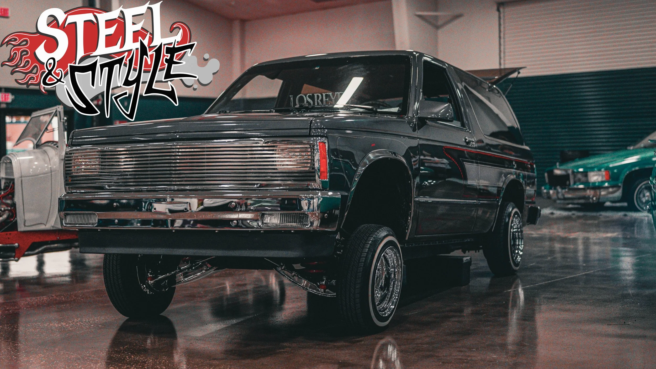 A black vintage van with chrome details and missing front wheels displayed in an indoor car showroom, with a logo that says 'Steel & Style' in the upper left corner.