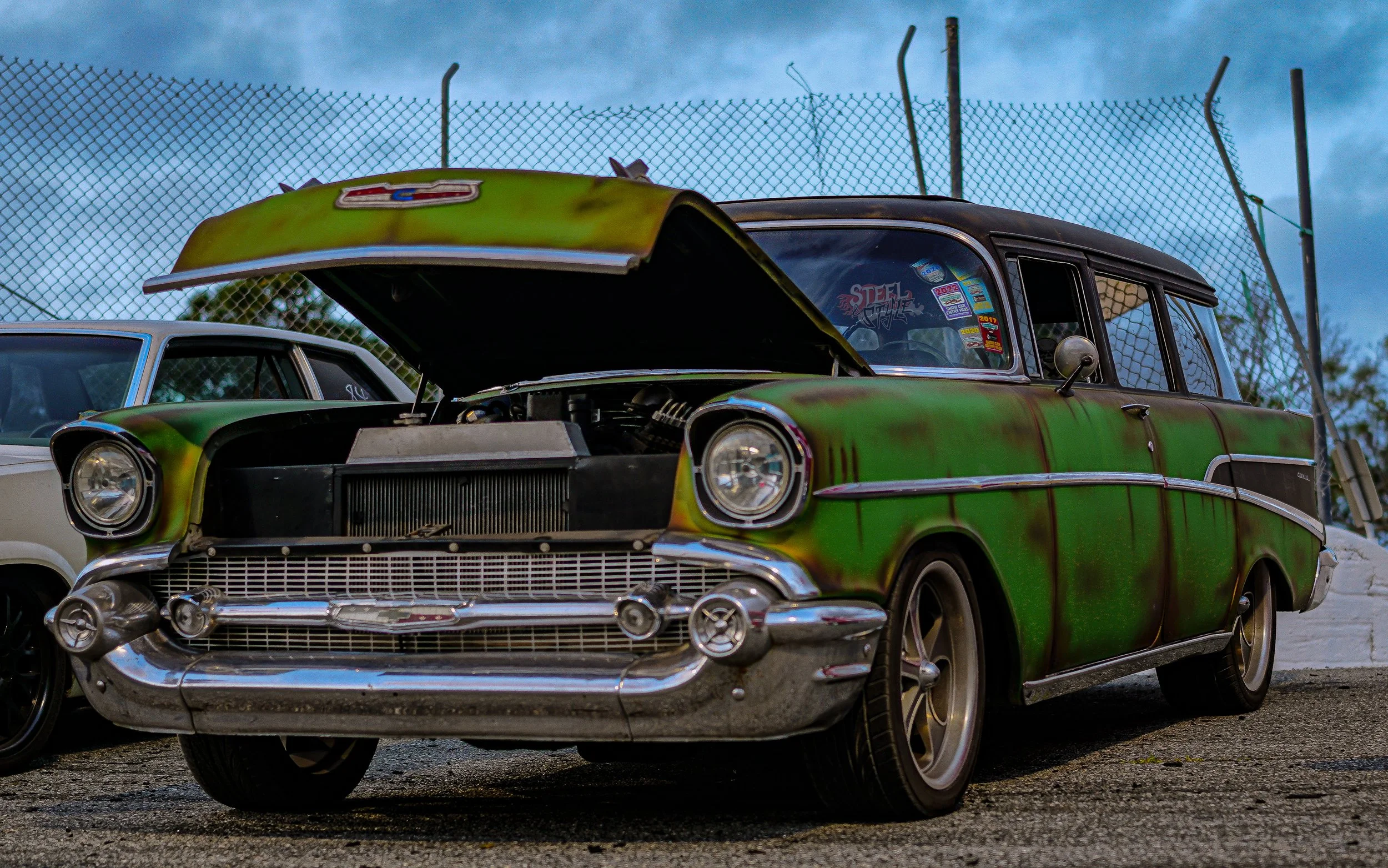 A vintage green and black station wagon with rust spots and a raised hood parked beside a white car at a car lot, with a chain-link fence in the background.