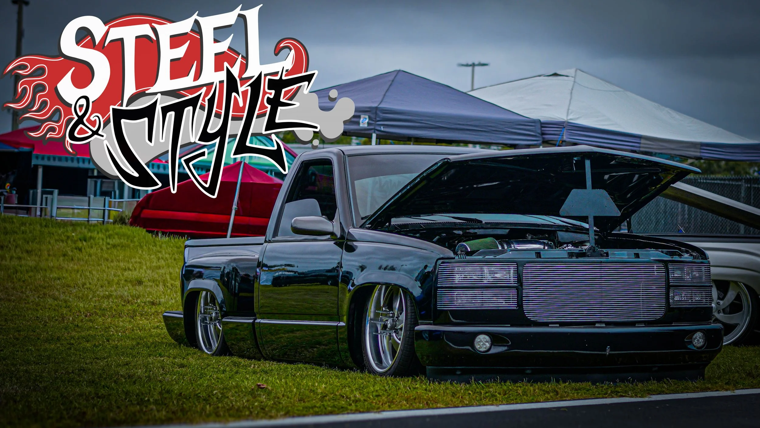 A heavily modified black pickup truck with an open hood, parked on grass at a car show, with tents and cloudy sky in the background, and the words 'Steel & Style' overlayed in graphic text.