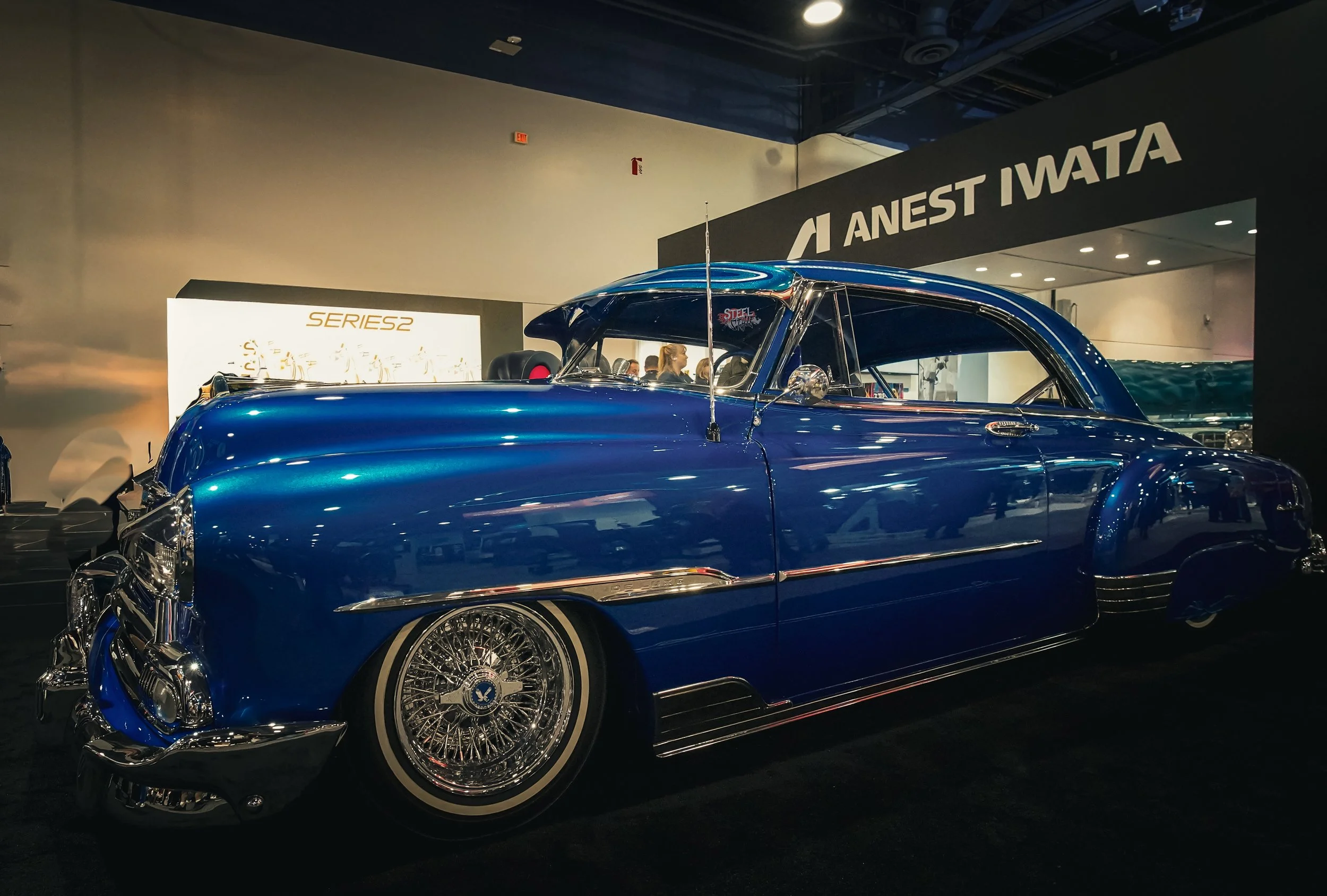 A vintage blue custom car on display at an auto show, with chrome detailing and wire wheels, positioned in front of signs that include 'Anest Iwata' and 'Series 2' visible in the background.