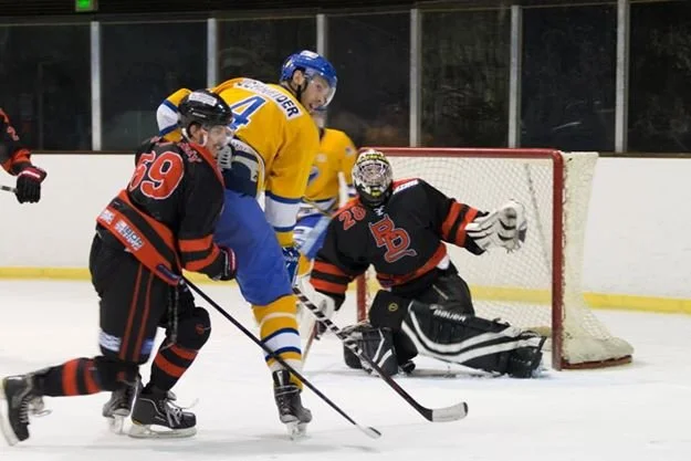 Hockey players in black and red jerseys try to score against a goalie in black and white gear, with a yellow-jersey player nearby, in an ice rink.