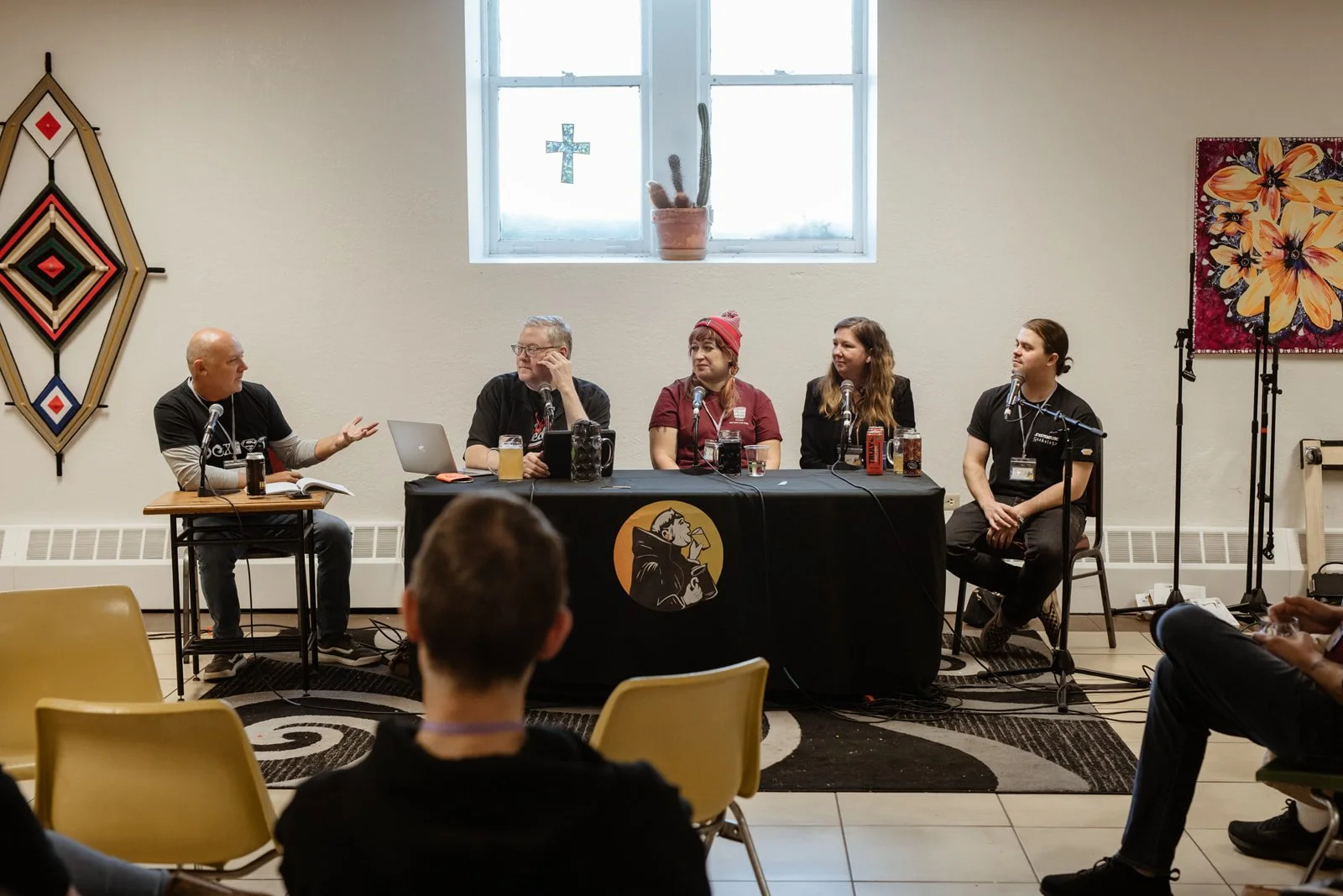 A panel discussion with five people seated at a table in front of an audience. The panel includes three men and two women, with microphones in front of them. The setting has a window, some artwork on the walls, and beers on the table.