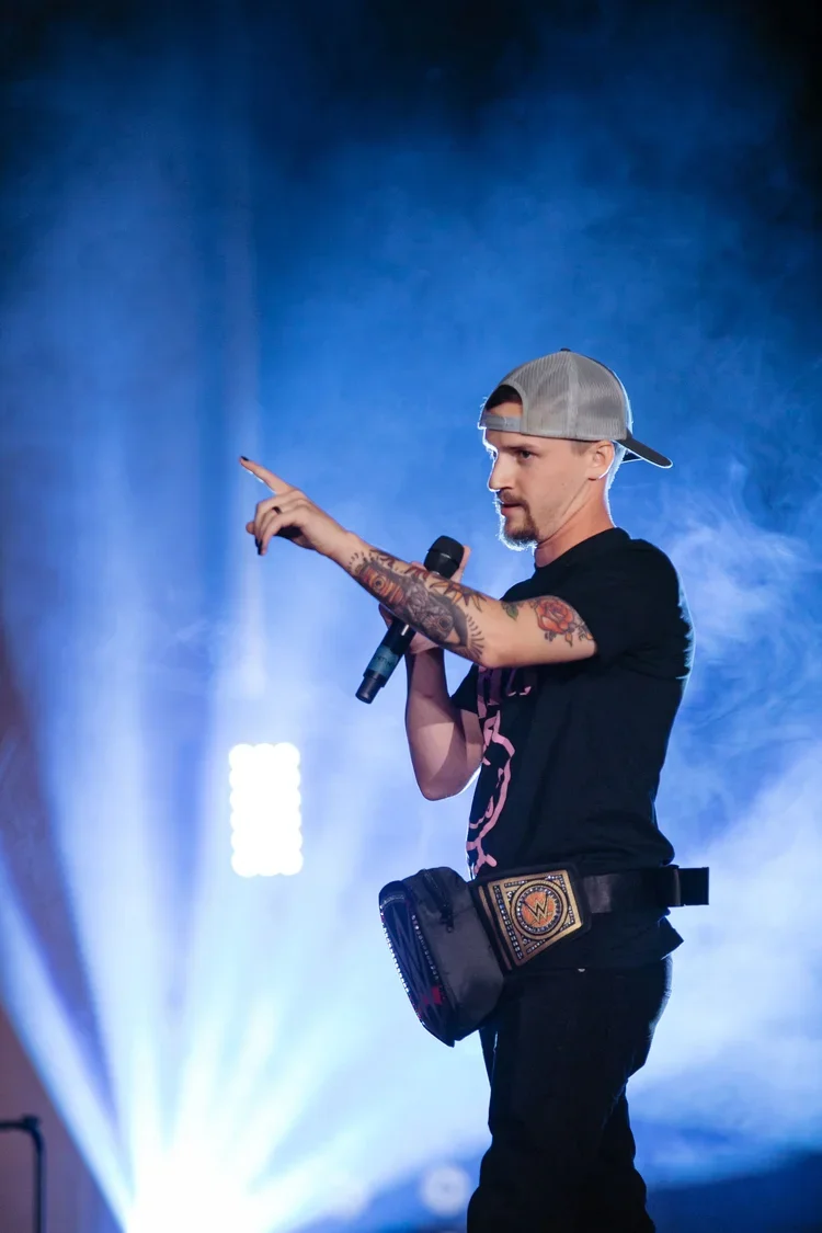 Male comedian performing on stage, holding a microphone, wearing a backward baseball cap, black t-shirt, and a fanny pack, with stage lights and smoke in the background.