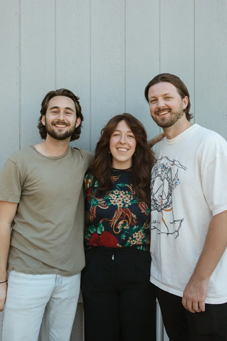 Three smiling young adults, two men and one woman, standing close together against a light gray wooden background.