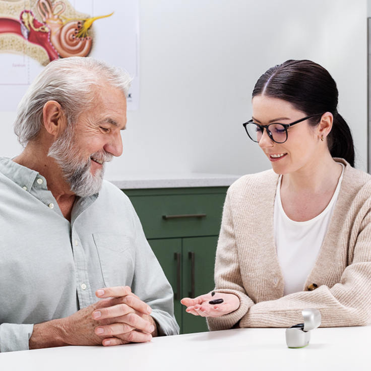 A nurse wearing a mask and a woman, sitting at a table with a computer, engaged in a conversation, possibly a consultation or interview.