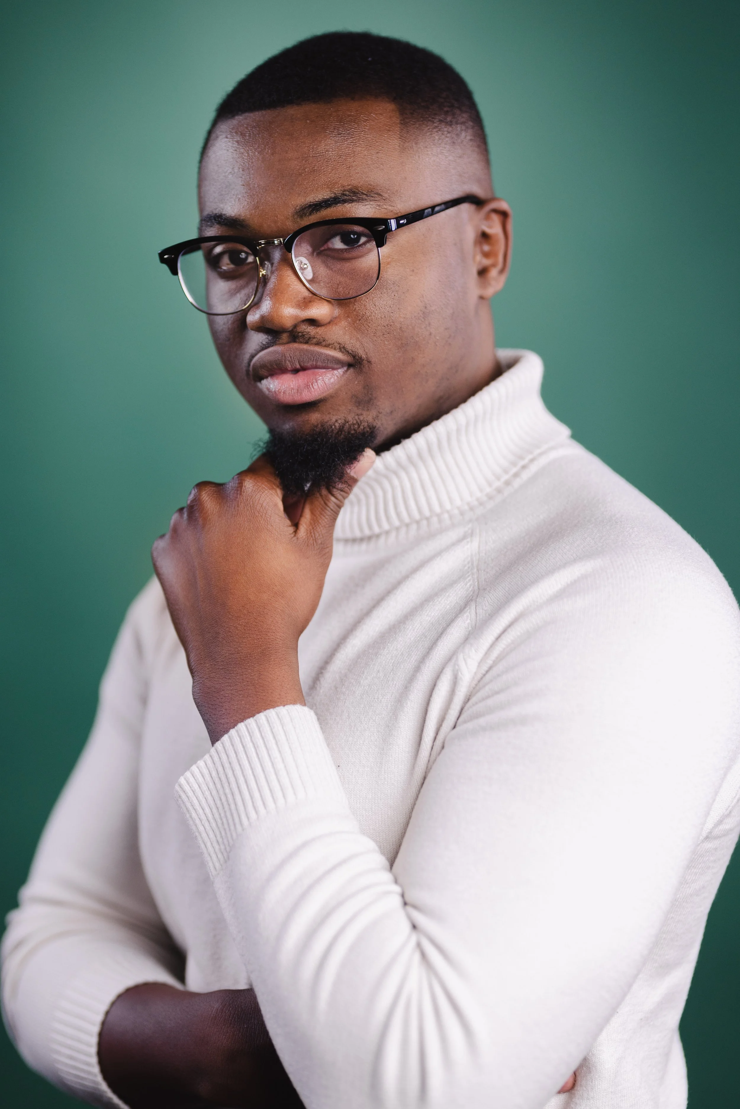 A man with glasses and a short beard wearing a white turtleneck sweater, posing with his hand on his chin against a green background.