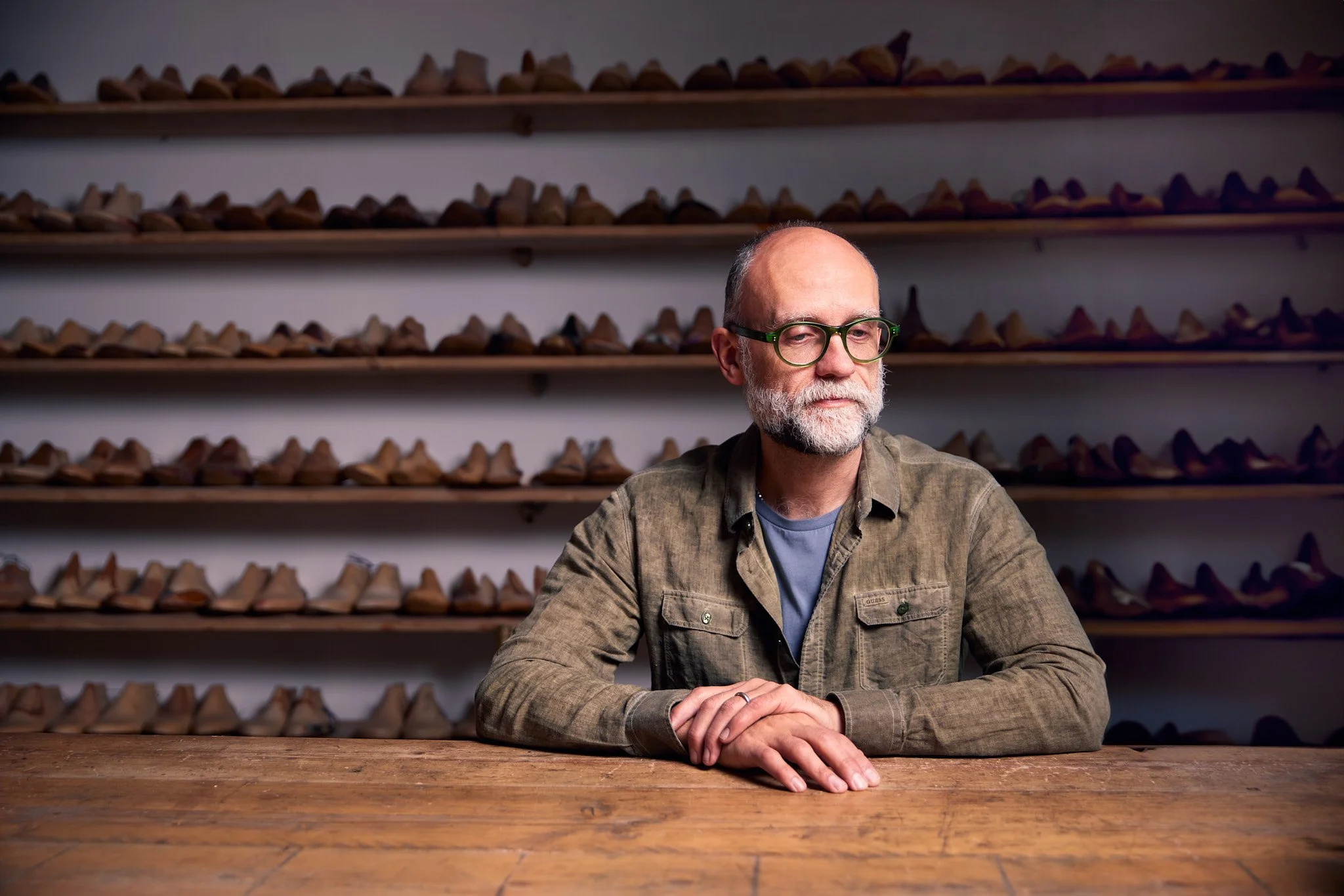 A man with glasses and a beard sitting at a wooden table in front of shelves filled with shoes.