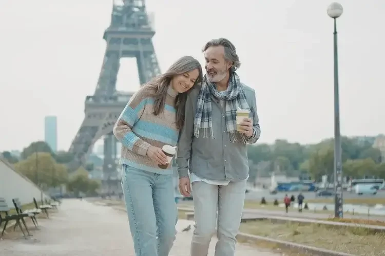 A middle-aged man and woman walking together with coffee cups in front of the Eiffel Tower in Paris, France.