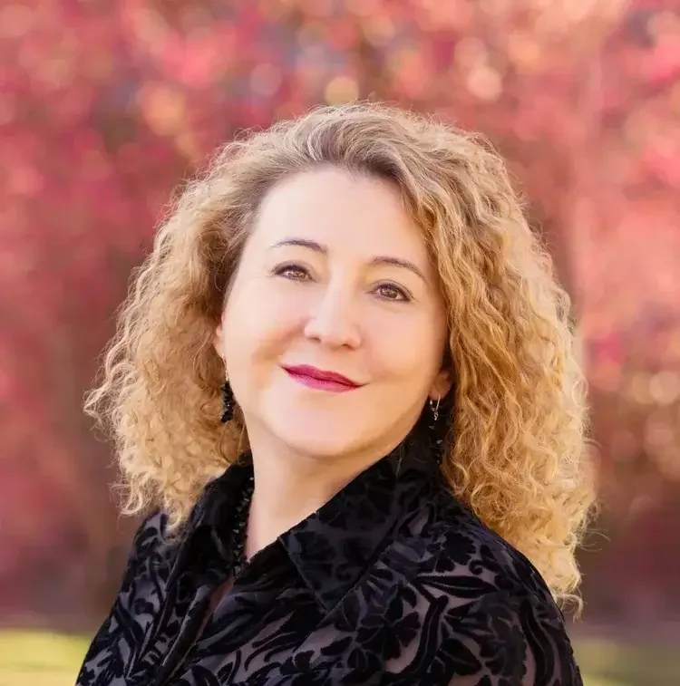 A woman with curly blonde hair smiling outdoors with a background of blurred pink and red foliage.