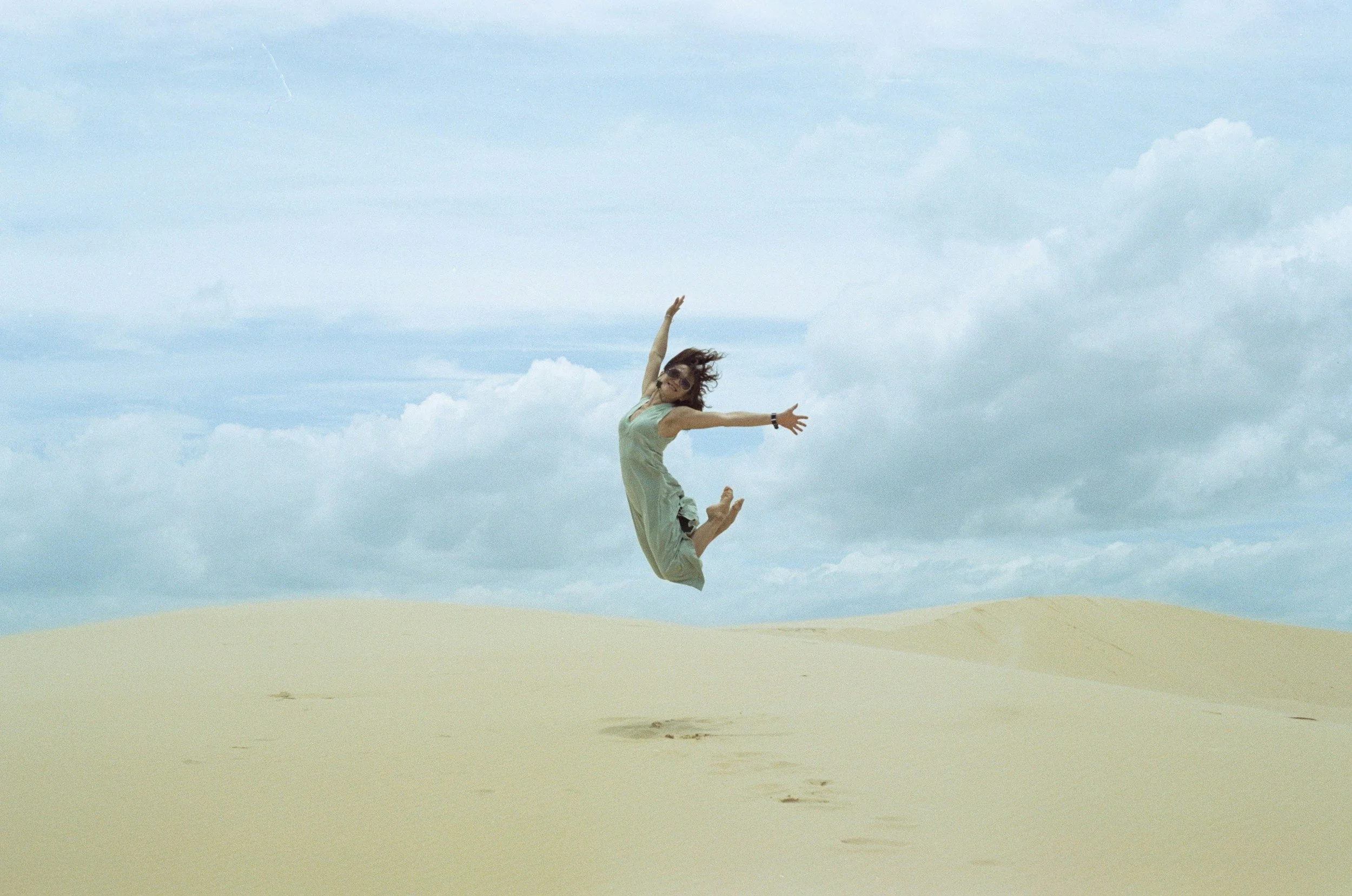 A woman in a dress jumping in the air on a sandy desert landscape with a cloudy sky in the background.