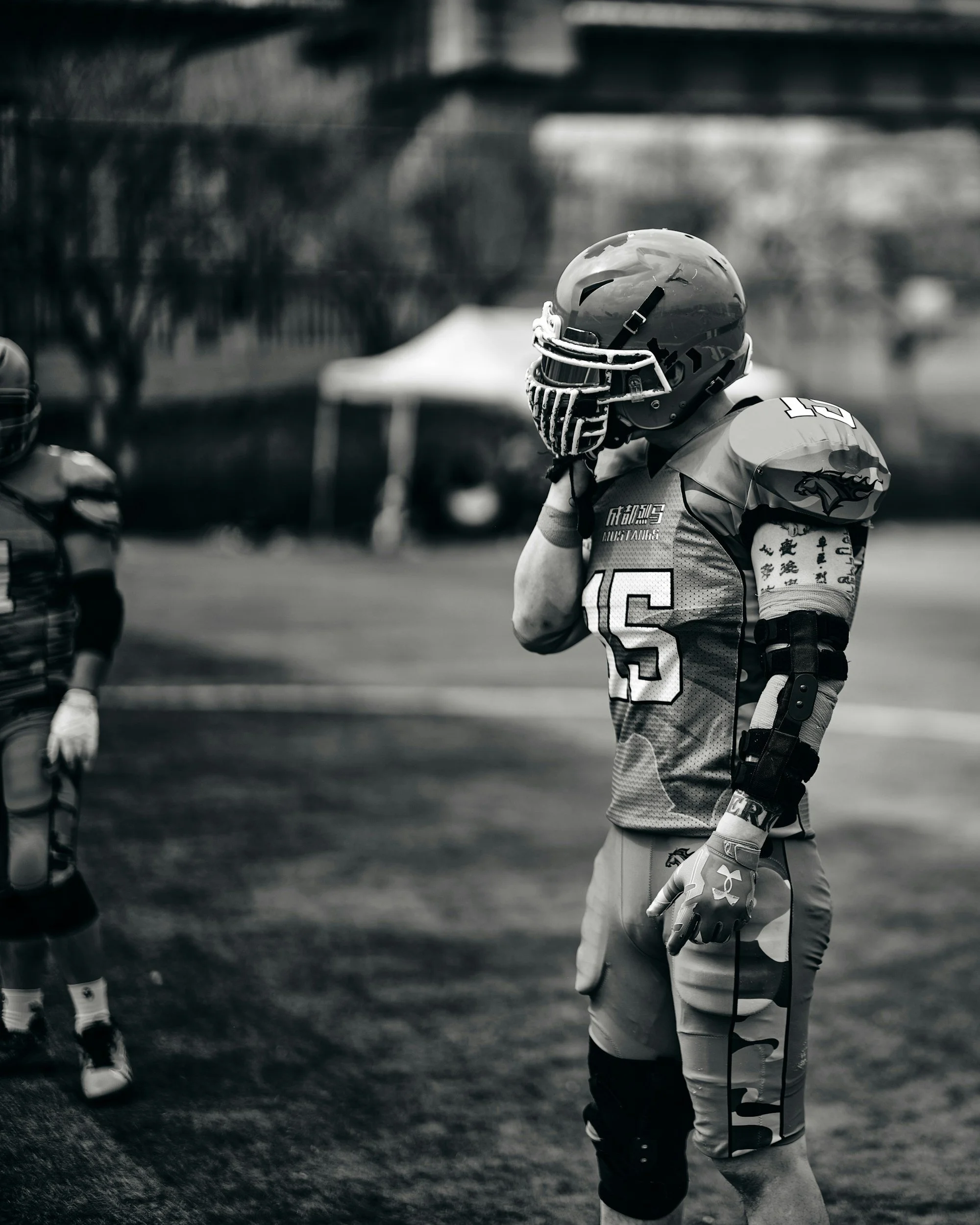 A football player wearing helmet, jersey, and pads stands on the field, touching his helmet, with another player partially visible on the side, and trees and a tent in the background.