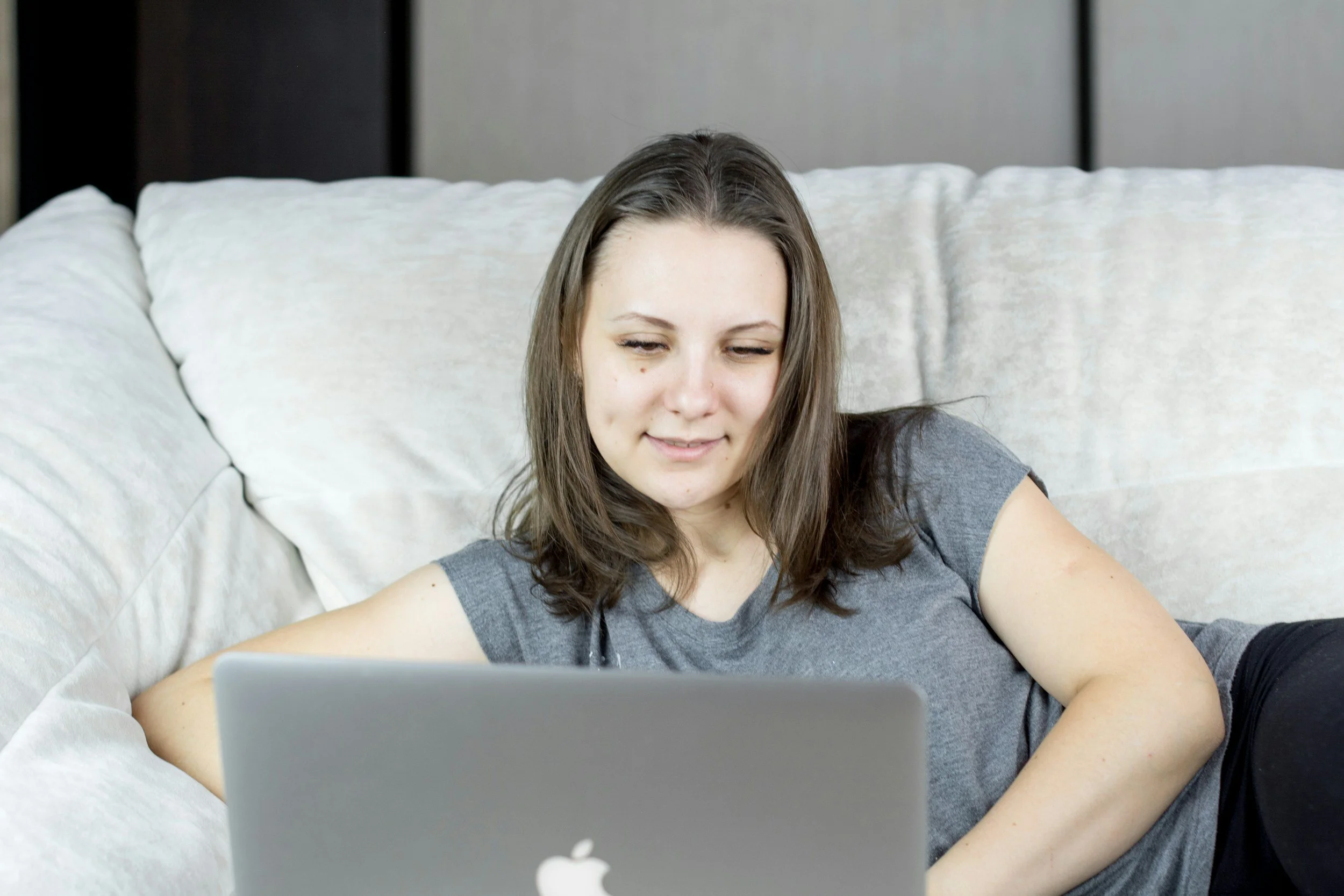 A woman with brown hair lying on a white couch with a gray shirt, looking at a silver MacBook laptop.
