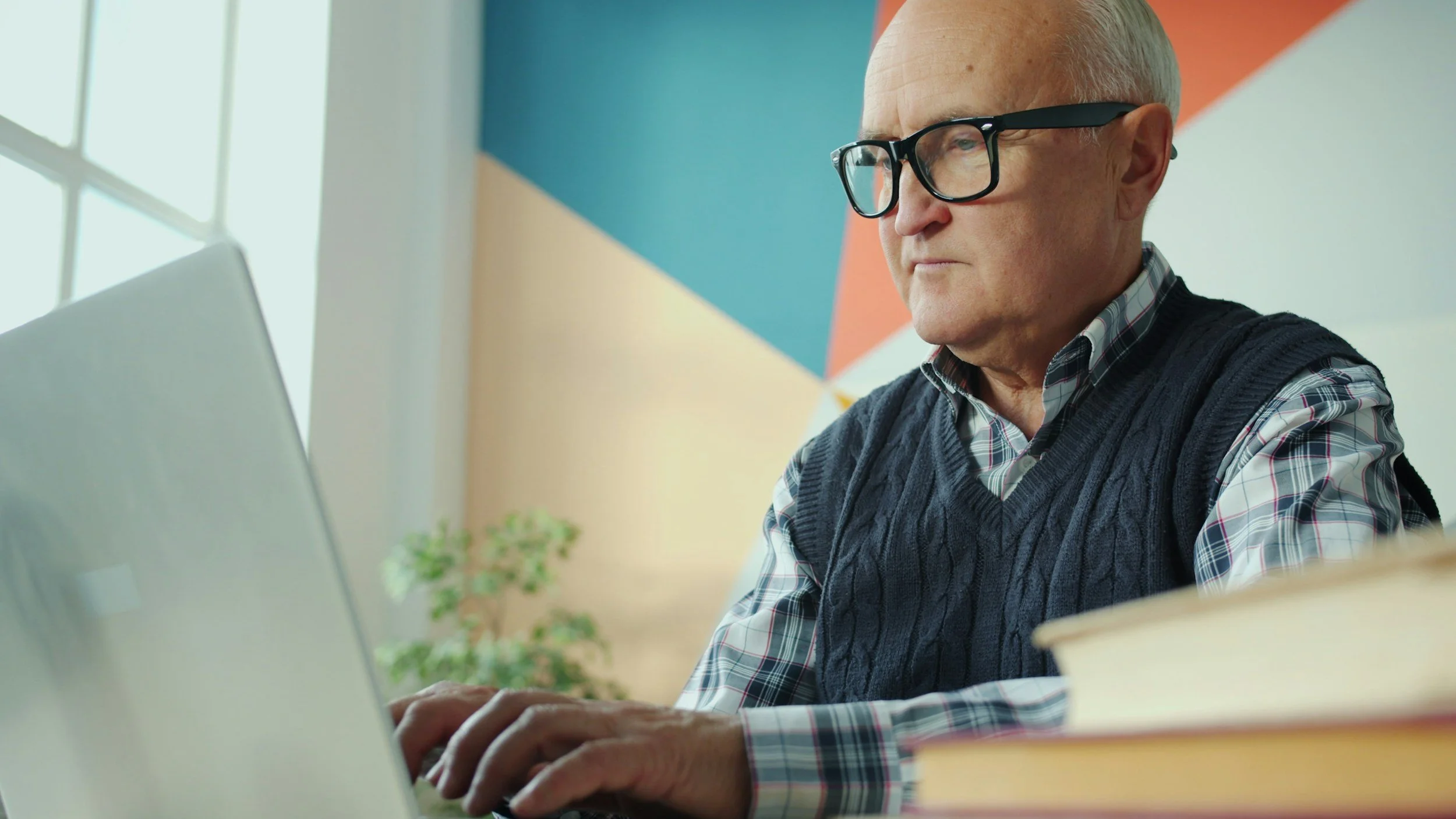 An elderly man with glasses working on a laptop at a desk, with books and colorful wall in the background.
