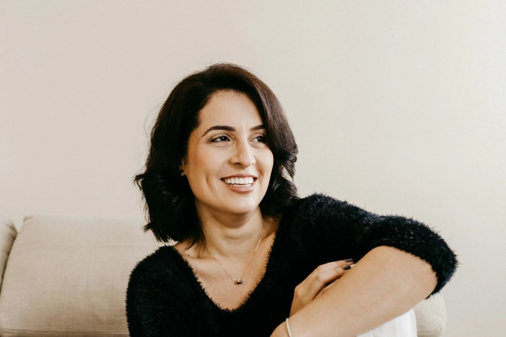 A woman with shoulder-length dark hair, smiling and sitting on a beige couch, wearing a black fuzzy top and a necklace.