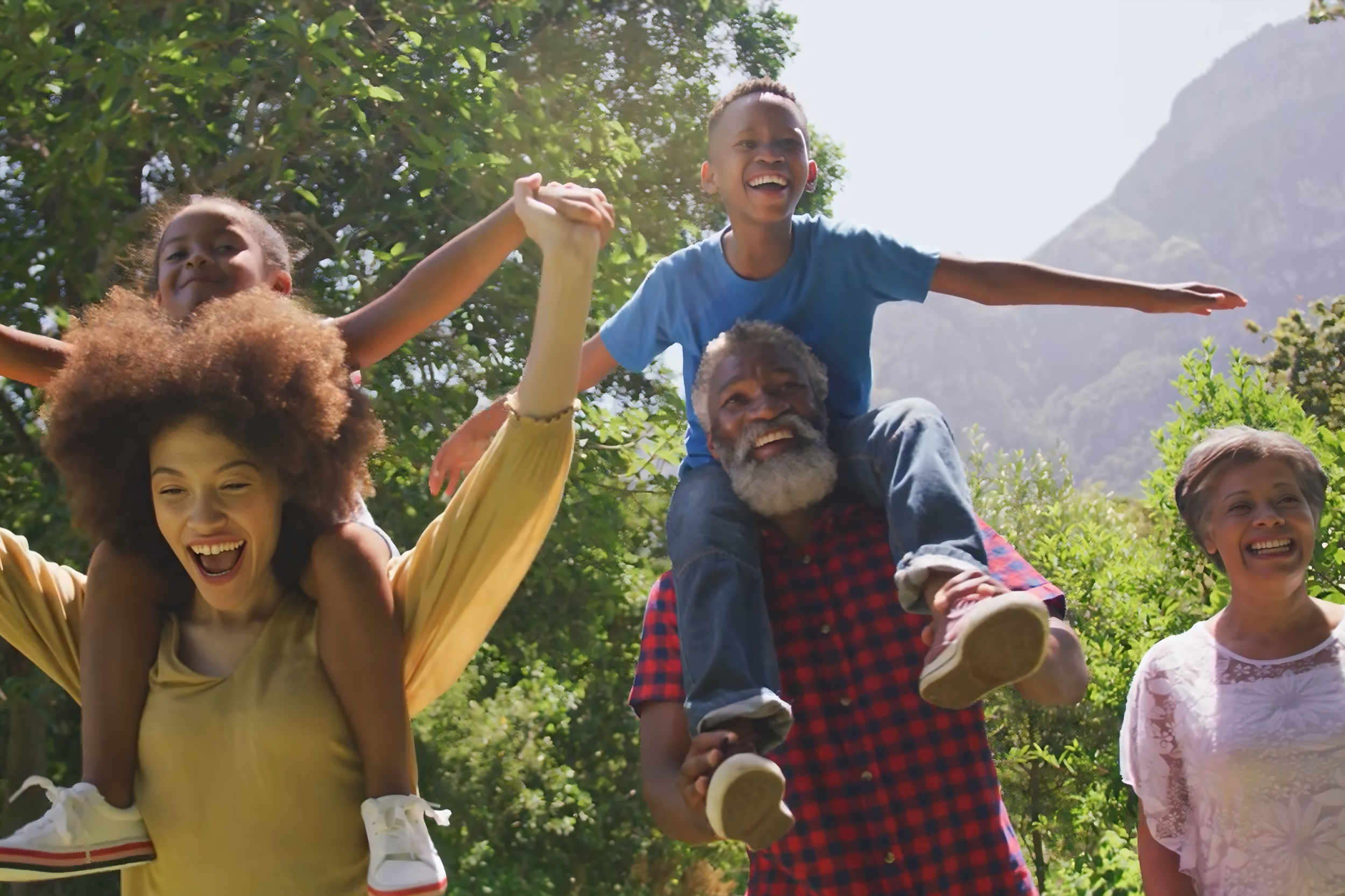 A group of diverse people smiling and enjoying a outdoor activity resembling a piggyback ride or a similar play activity in a lush green park with trees and mountains in the background.