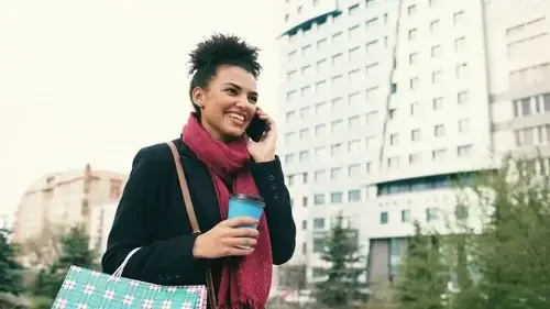 Woman smiling and talking on cell phone while holding a coffee cup, outdoors in an urban setting.