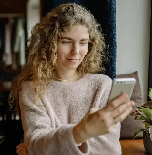 A young woman with curly blonde hair wearing a pink sweater looks at her smartphone while sitting indoors near a window.