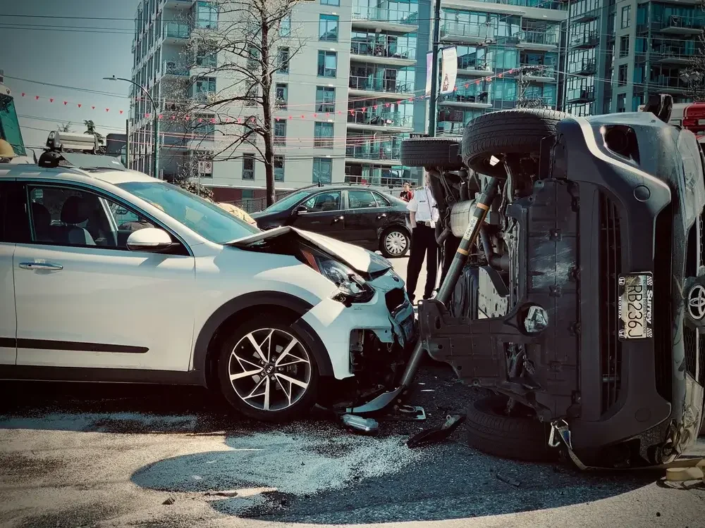 White car involved in an accident, collided with a black truck that is flipped onto its side, with visible damage and debris on the road, in an urban area with high-rise buildings.