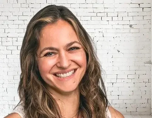 A smiling woman with wavy brown hair stands against a white brick wall.
