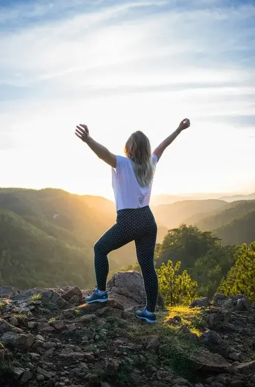 A woman standing on rocks with arms raised, overlooking a mountainous landscape during sunset.