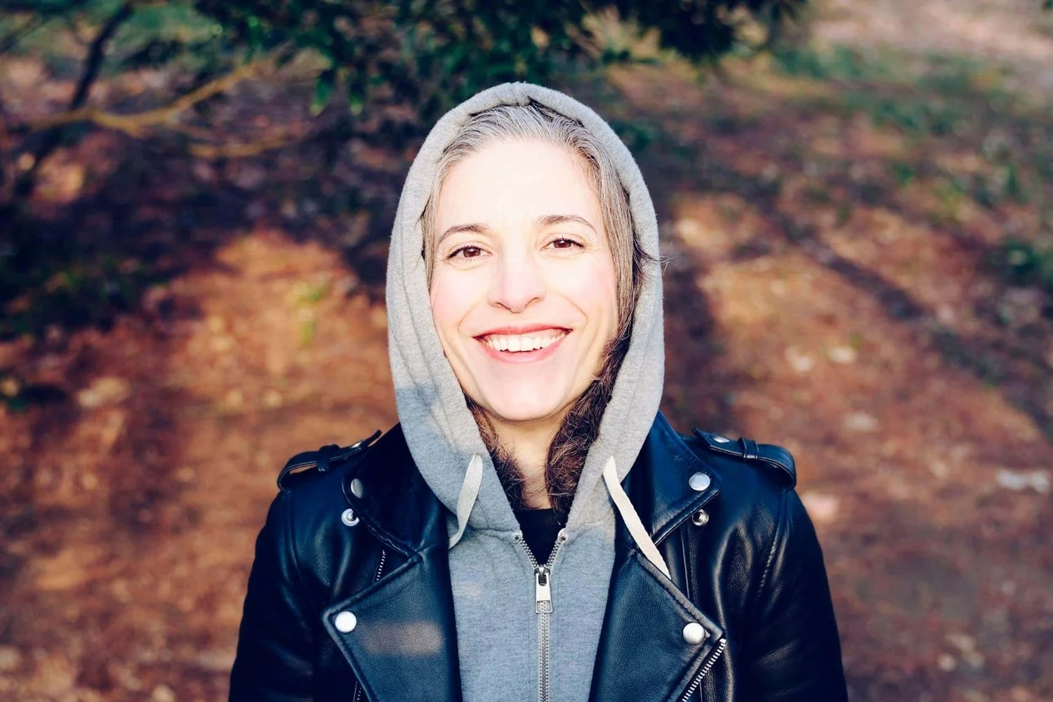 Smiling woman wearing a gray hoodie and black leather jacket outdoors with a blurred natural background.