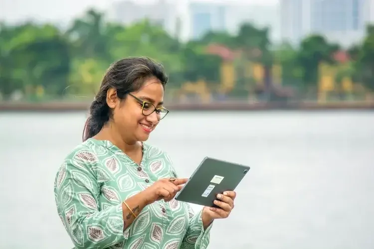 Woman with glasses and patterned blouse smiling while using a tablet outdoors near a body of water with trees and buildings in the background.