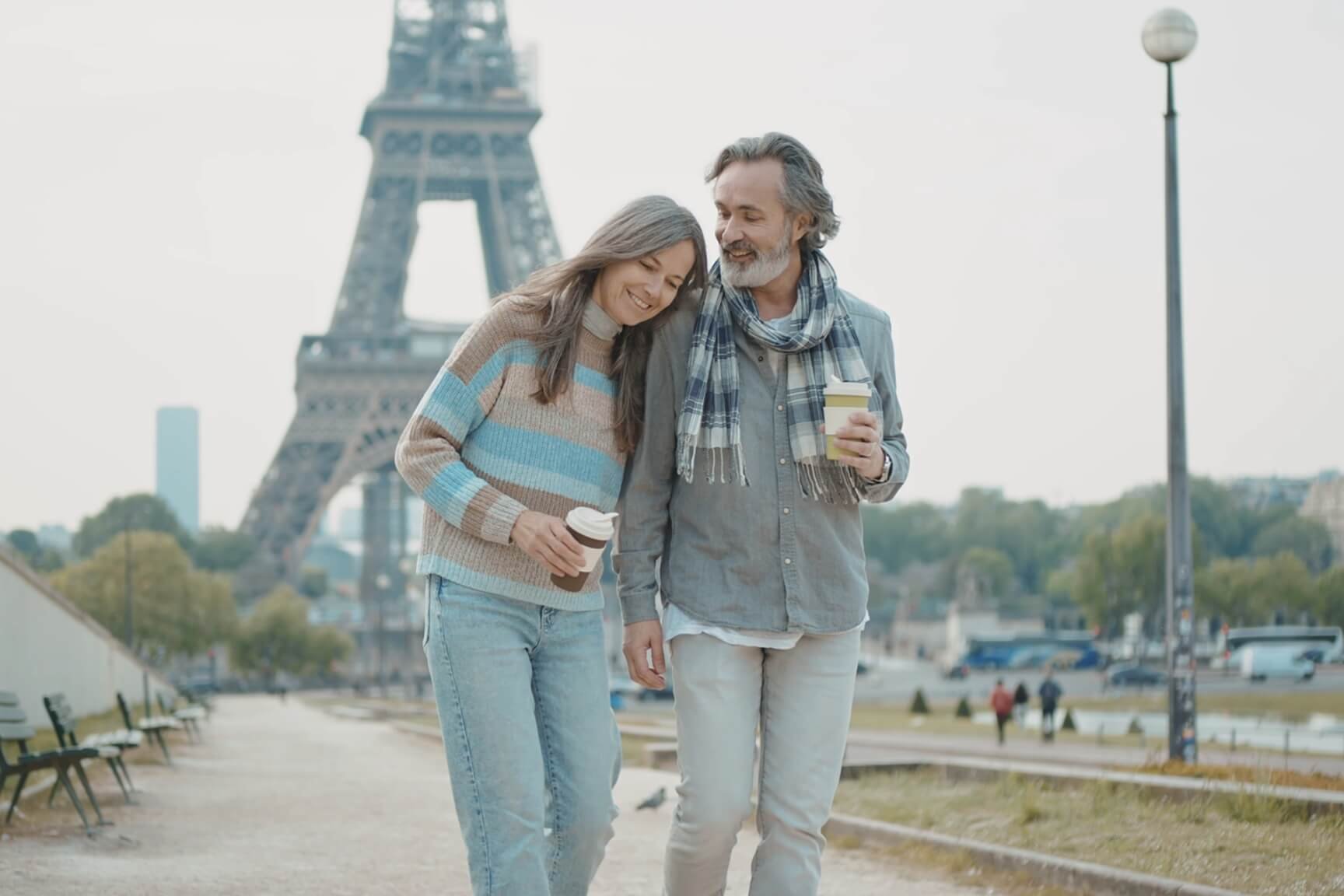 A couple strolls hand-in-hand in front of the Eiffel Tower, surrounded by lush greenery and a clear blue sky.