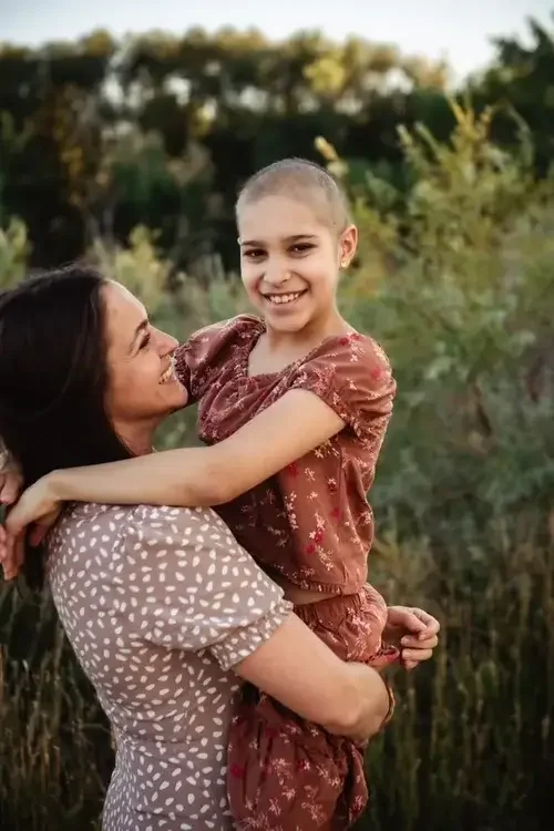 A woman holding a smiling girl with a shaved head outdoors in a natural setting.
