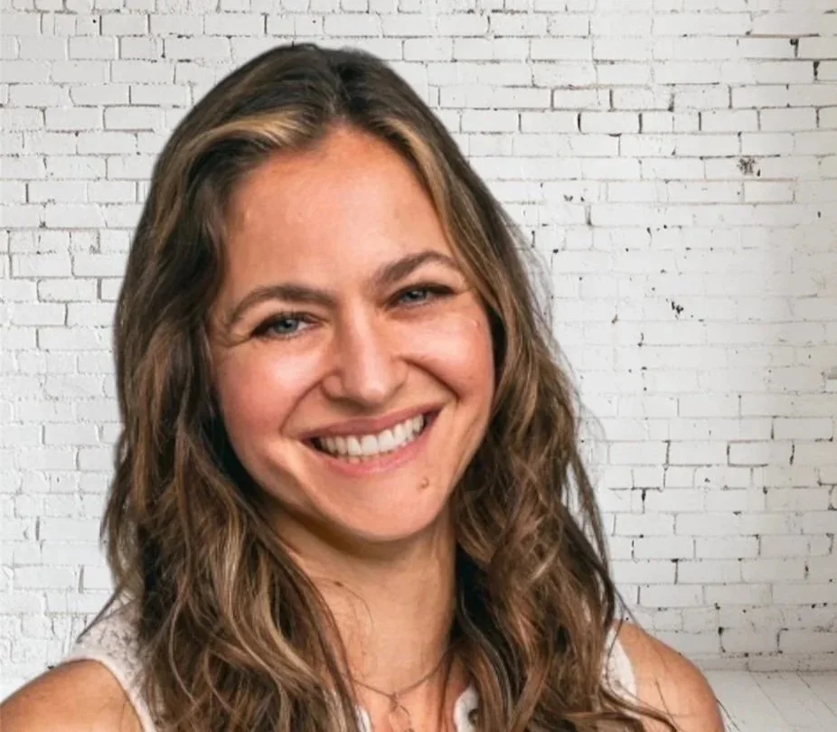 A woman with long, wavy brown hair, smiling in front of a white brick wall.