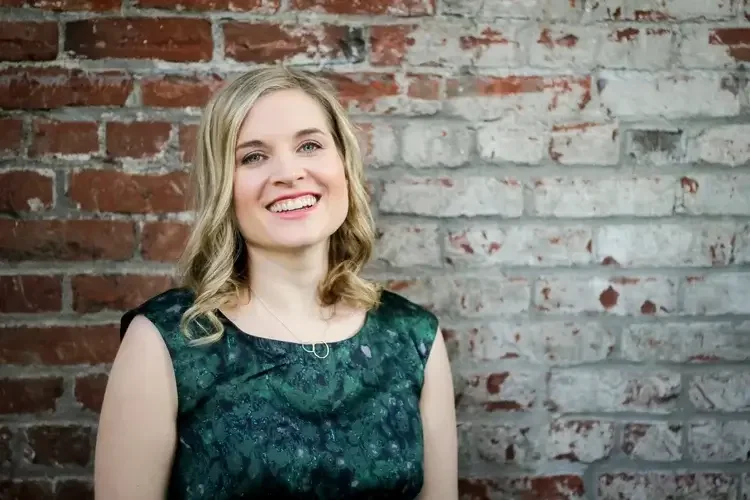A young woman with blonde hair and a big smile standing in front of a red brick wall.