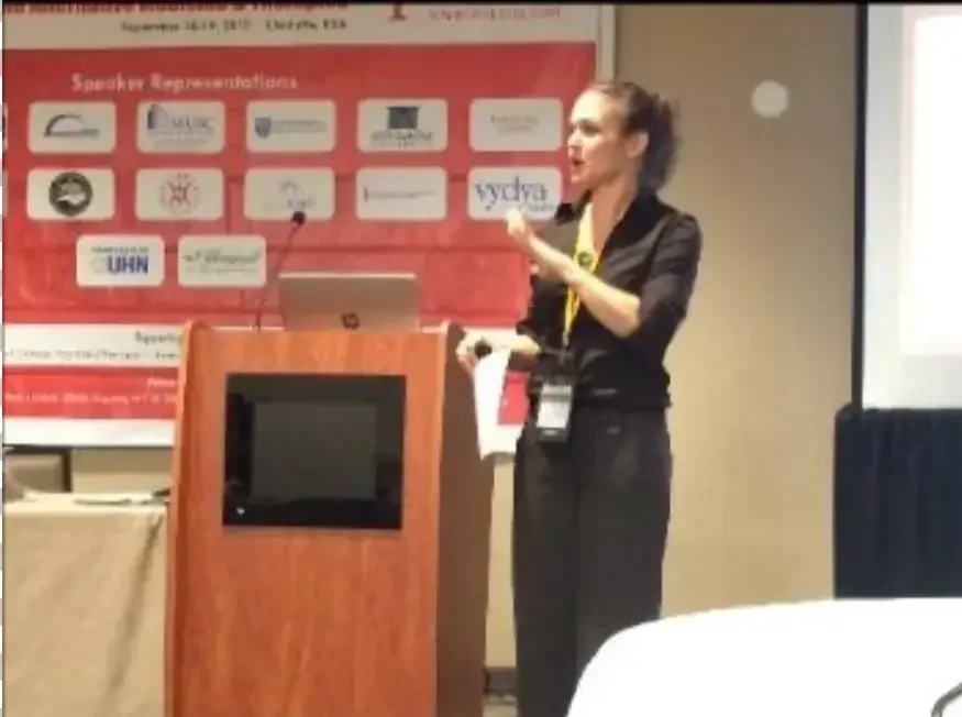 A woman giving a presentation at a conference, standing behind a wooden podium with a laptop. There is a speaker's banner with logos and text in the background, and a projection screen to her right.