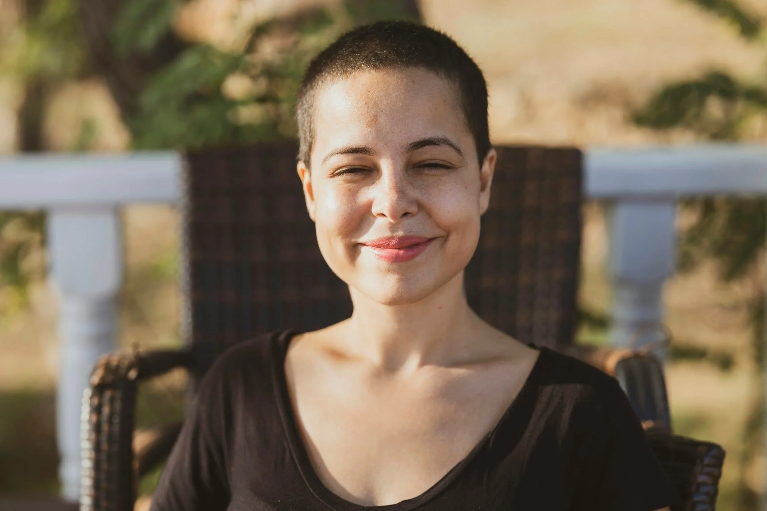 A young woman with a shaved head smiling outdoors, sitting on a patio chair with sunlight on her face.
