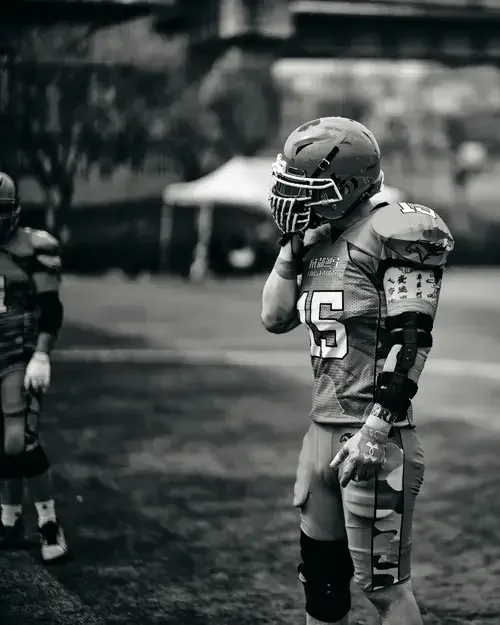 A young football player standing on the field, wearing a helmet, jersey with the number 16, and protective gear, in a moment of reflection or concern.