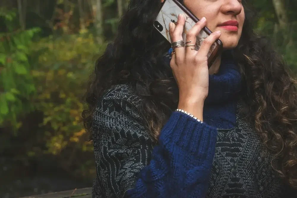 A woman with curly hair, wearing a patterned jacket and a blue sweater, talking on a smartphone outdoors.