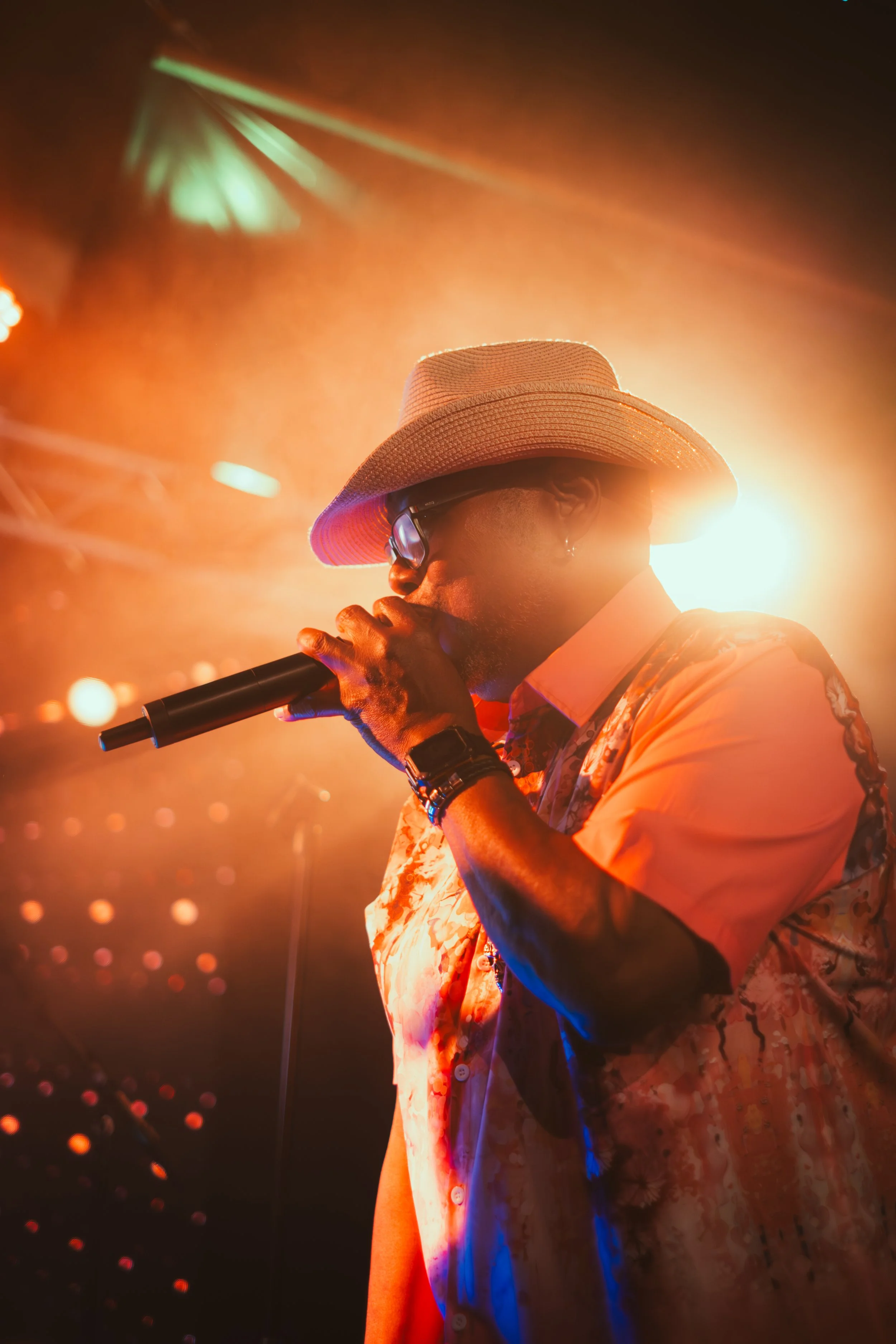 Ein Musiker trägt einen Strohhut und eine Sonnenbrille, singt in ein Mikrofon, hinter ihm leuchten bunte Bühnenlichter bei einem Auftritt im Dunkeln.