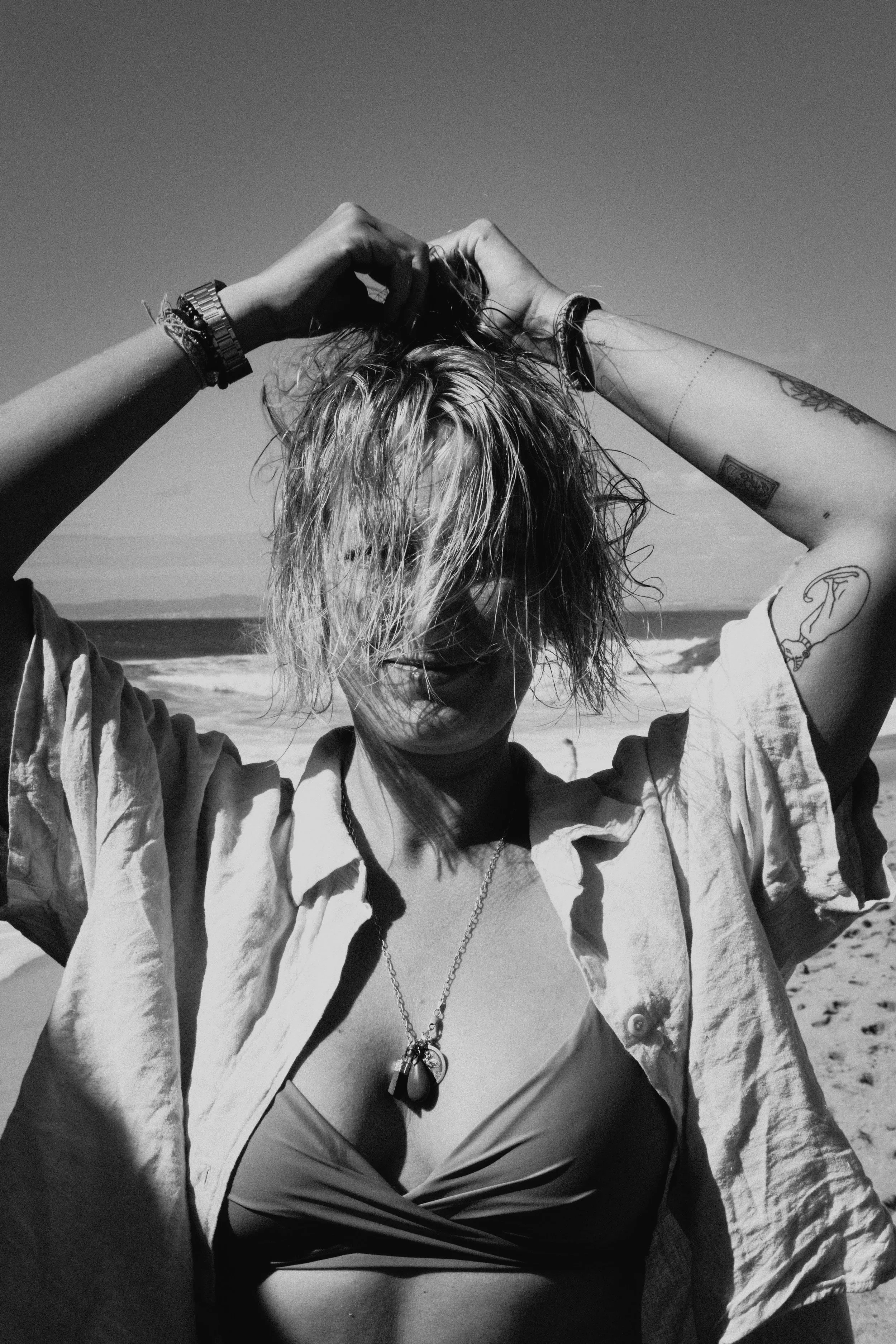 Junge Frau mit nassem, zerzaustem Haar am Strand, trägt Bikini und offene Hemd, macht eine Pose mit erhobenen Armen, schwarze und weiße Farben, Tattoos, Sonnenbrille auf dem Kopf.