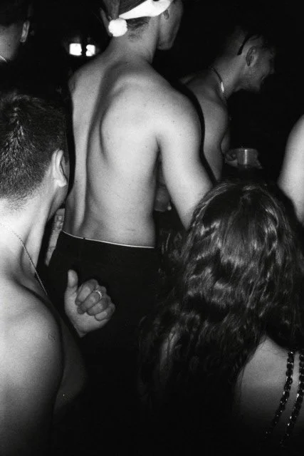 Gruppe junger Menschen bei einer Party, einige nackt oder nur mit Unterwäsche, in einer dunklen Umgebung, vermutlich in einem Club oder einer Feier.