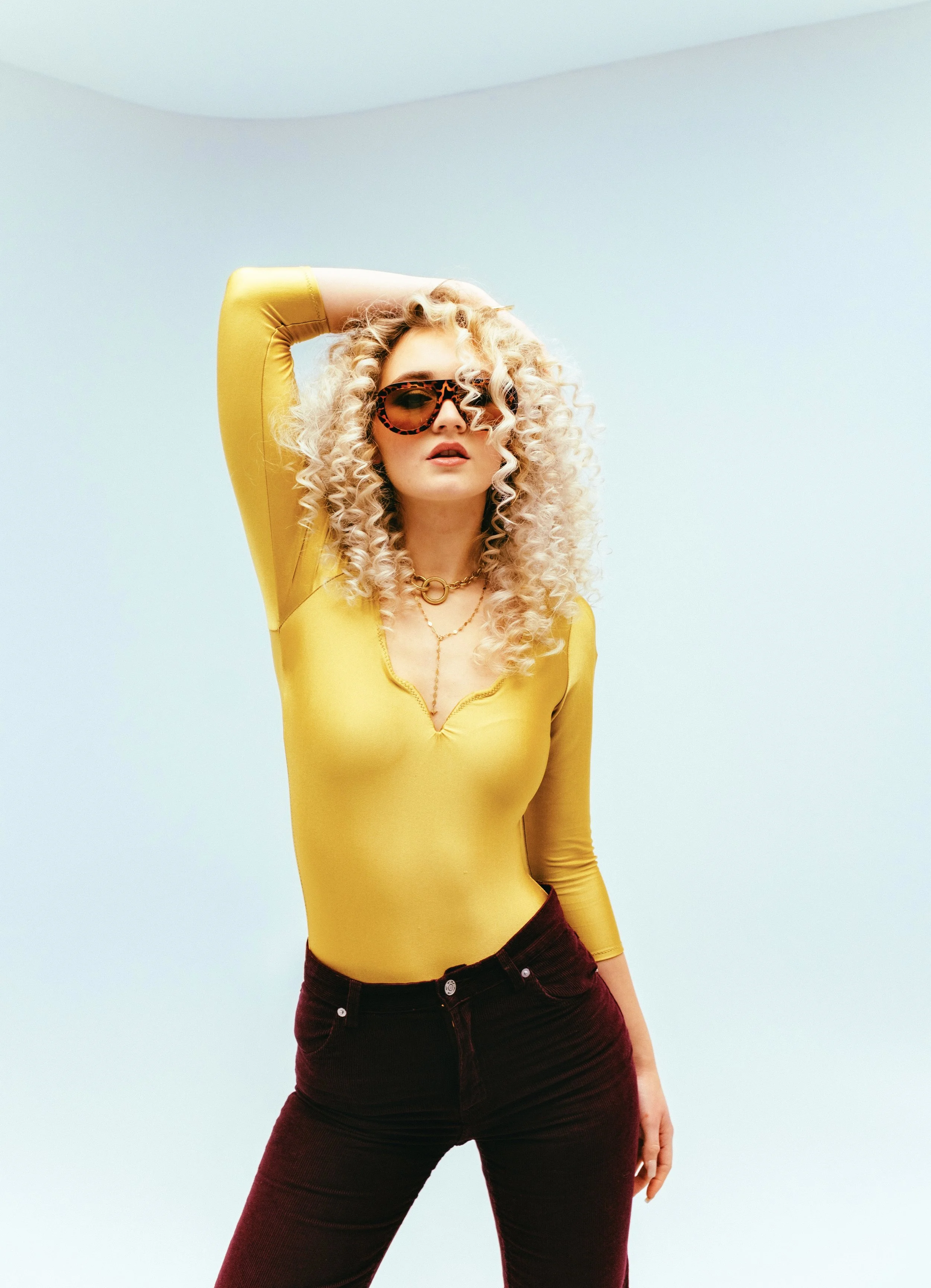 Junge Frau mit lockigem blondem Haar, Sonnenbrille im Leopardenmuster, gelber Langarm-Body und roten Hosen posiert vor hellblauem Hintergrund.