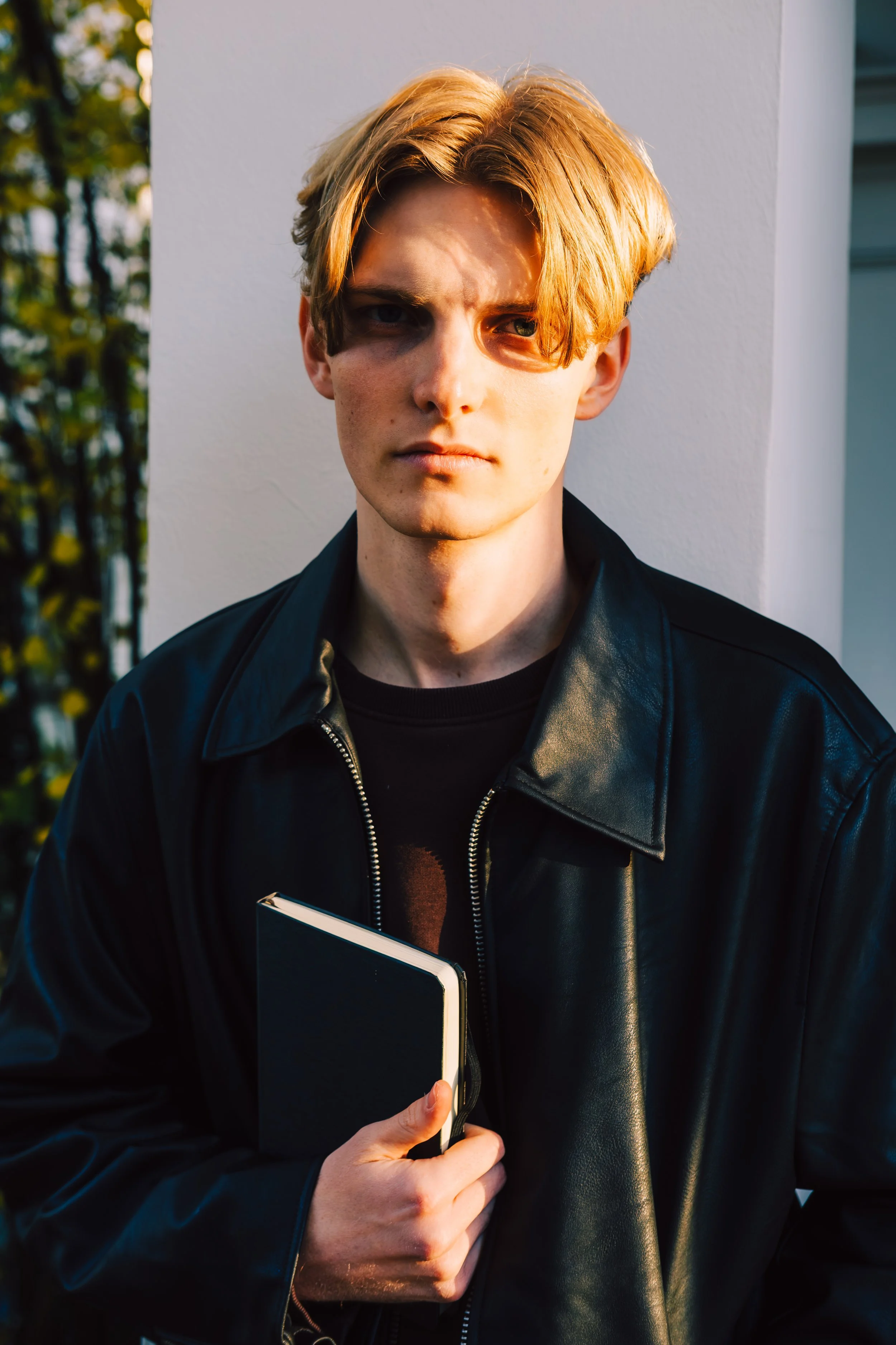 Junge Person mit blonden Haaren, trägt schwarze Lederjacke, hält ein schwarzes Notizbuch, steht draußen bei Sonnenuntergang