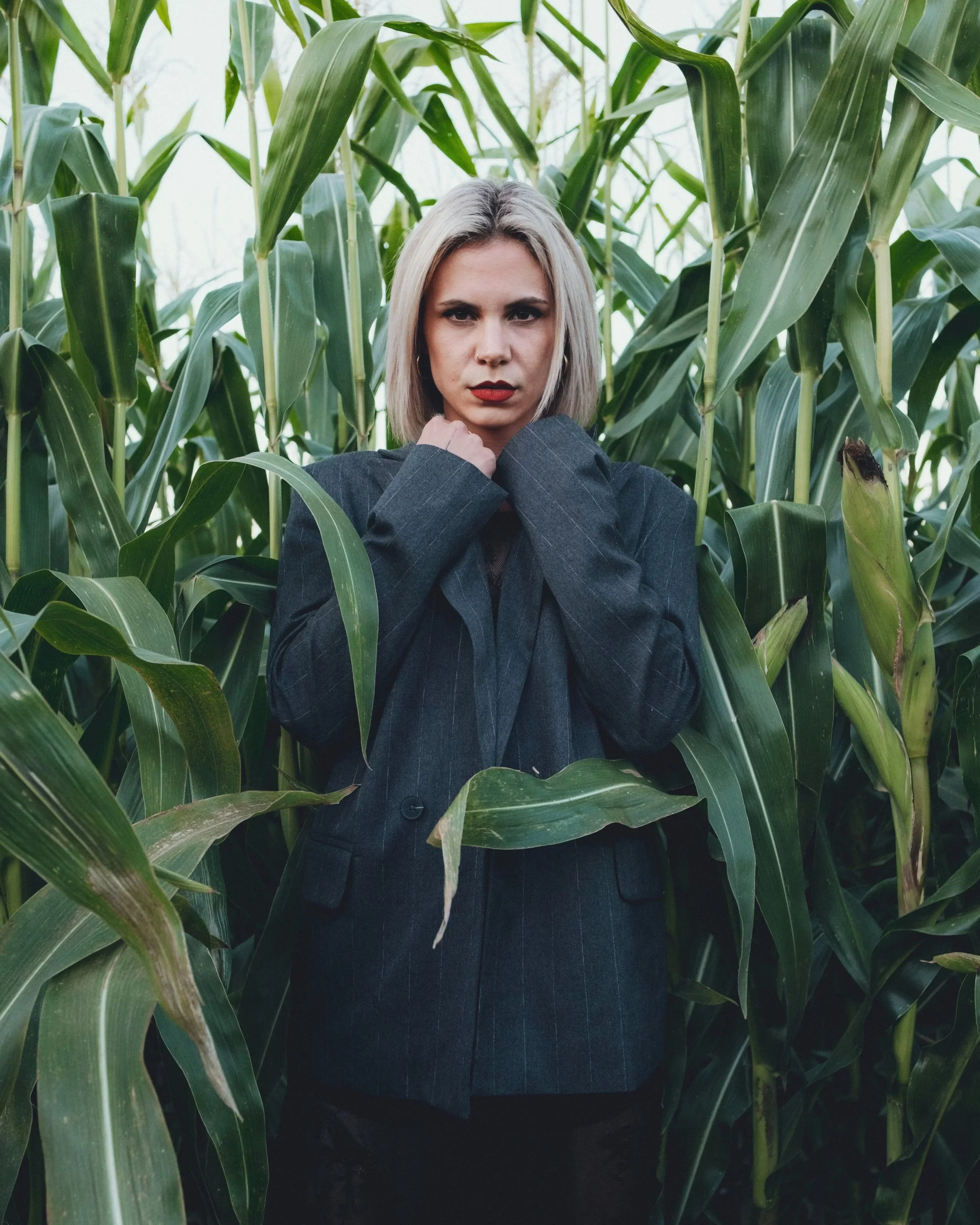 Eine Frau mit blondem, kinnlangem Haar trägt eine dunkle Anzugjacke und steht in einem Feld mit hohen Maispflanzen, während sie direkt in die Kamera schaut.