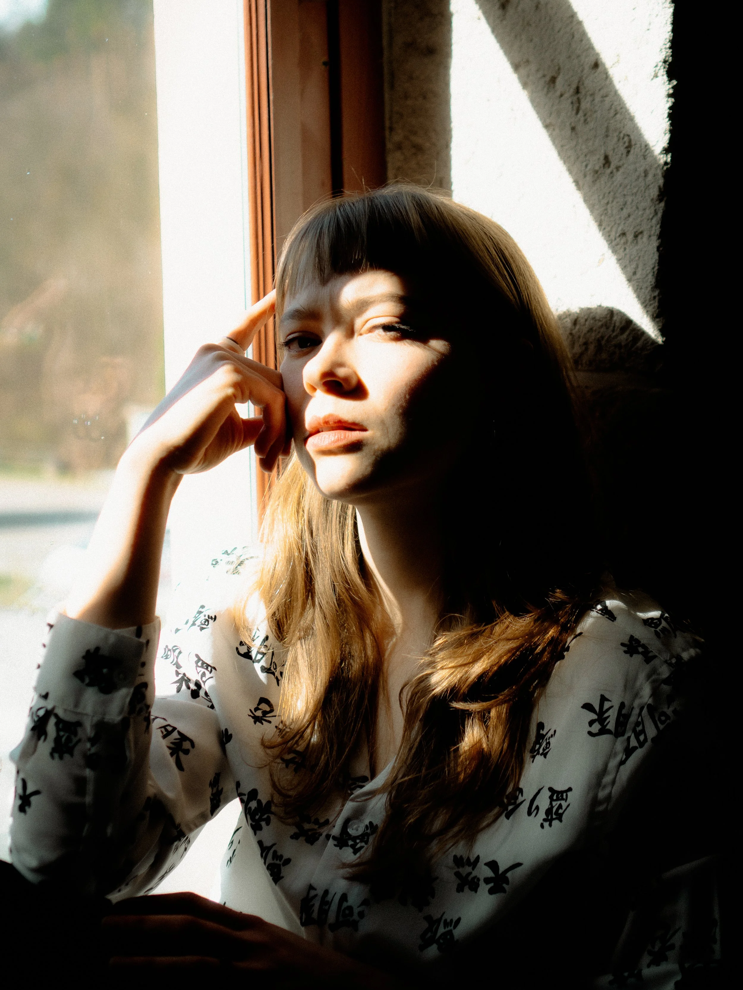 Nahaufnahme einer jungen Frau, die an einem sonnigen Tag am Fenster sitzt, mit Sonnenlicht auf ihrem Gesicht, trägt eine weiße Bluse mit schwarzen Mustern, blickt nachdenklich.