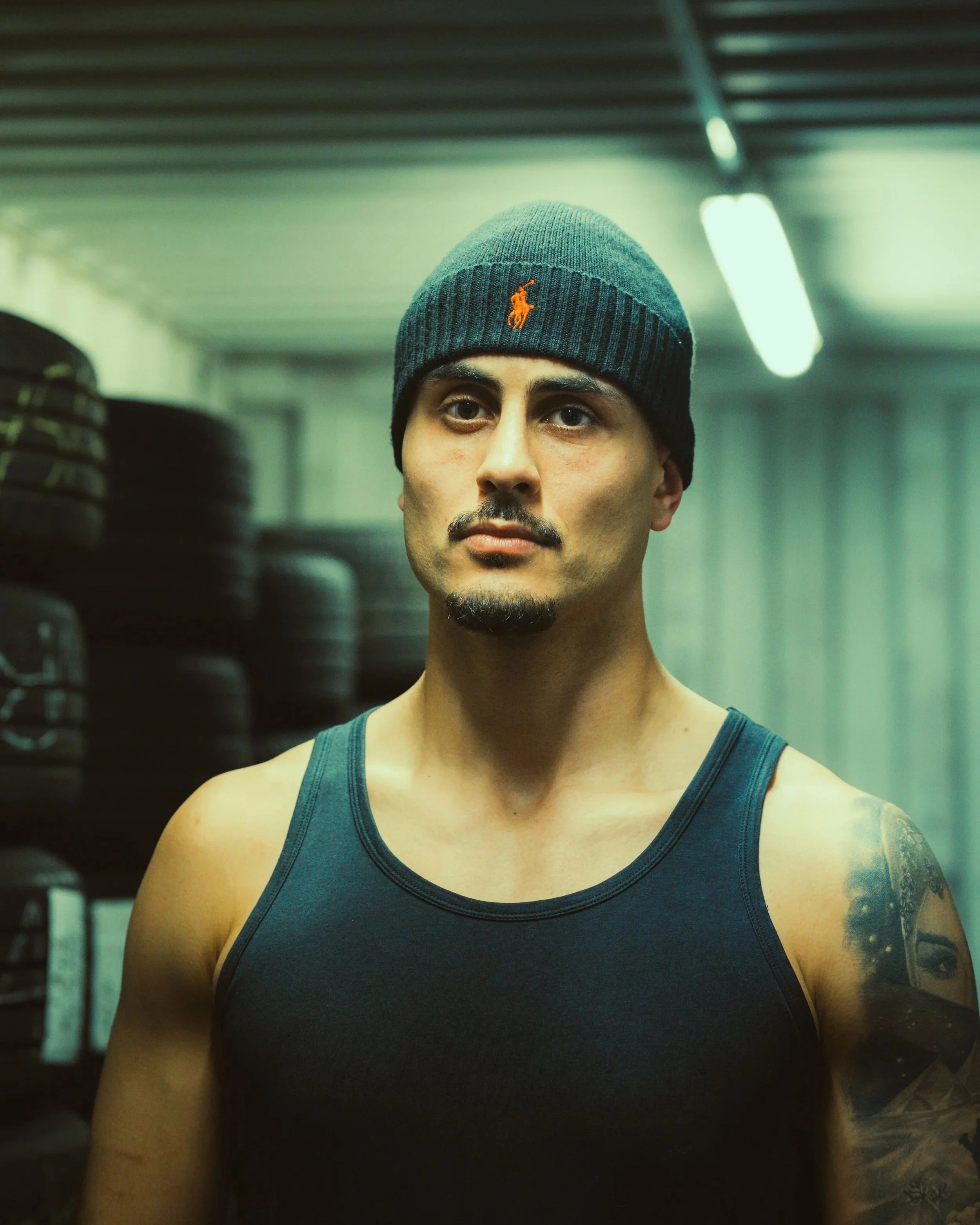 Mann in Trainerjacke mit Tattoo am Arm steht in einem Fitnessstudio vor Regal mit Reifen