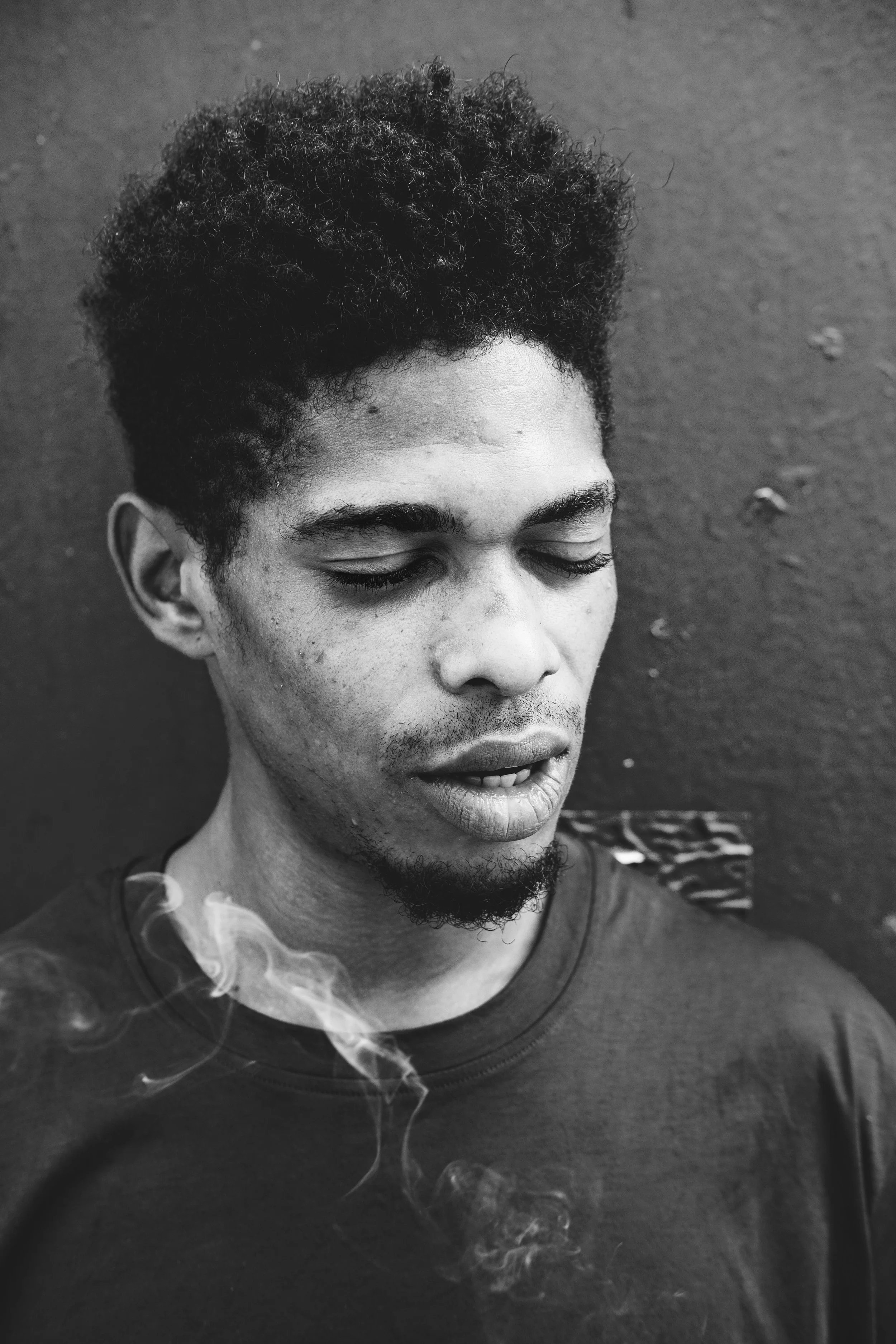 Schwarz-weiß Portrait eines jungen Mannes mit offenen Augen, dunklen, krausen Haaren und leichtem Bart, der eine Zigarette raucht, Rauch steigt auf.