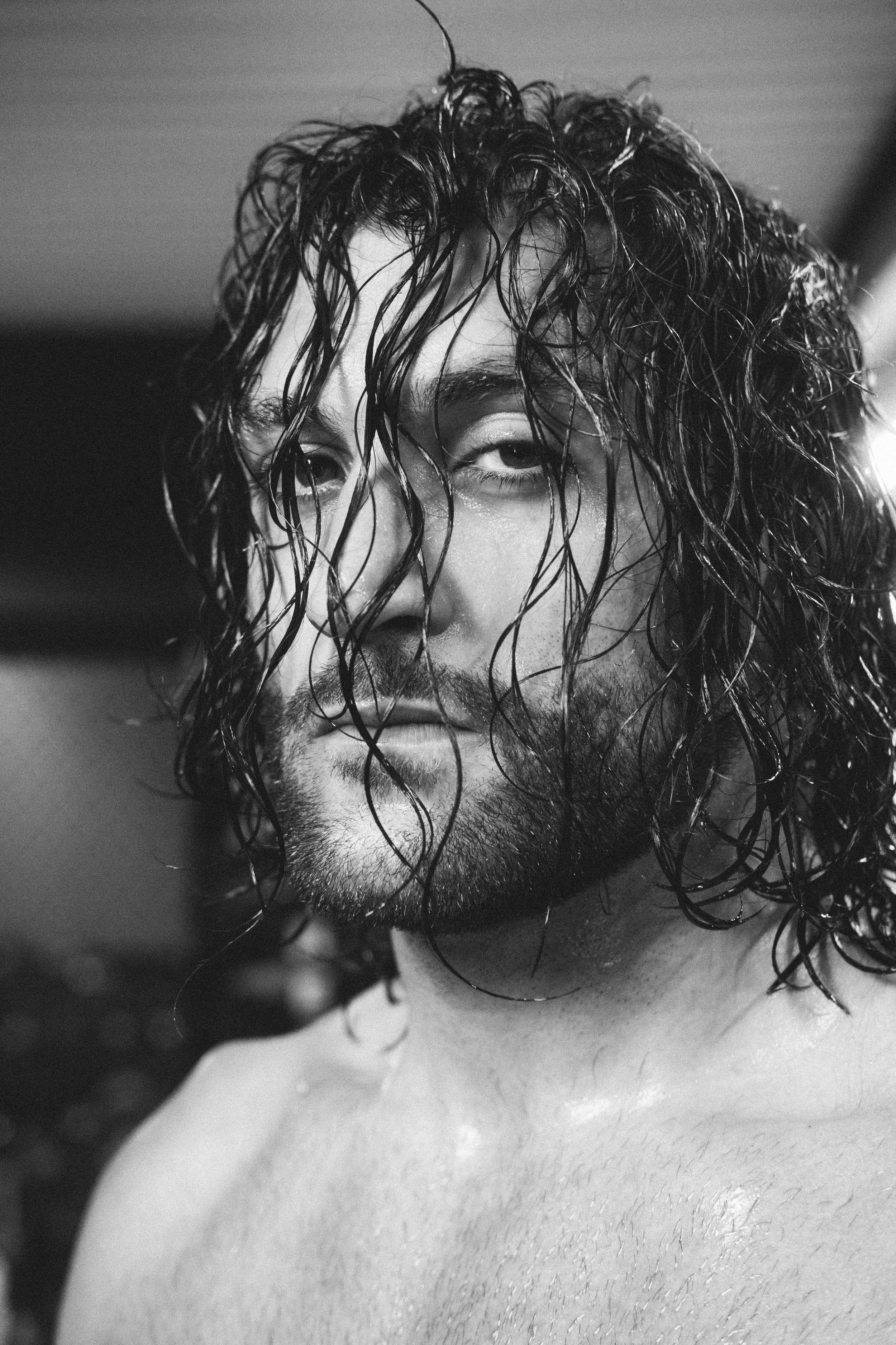 Schwarz-weiß Porträt eines Mannes mit nassen, lockigen Haaren und Bart, der die Kamera direkt ansieht.